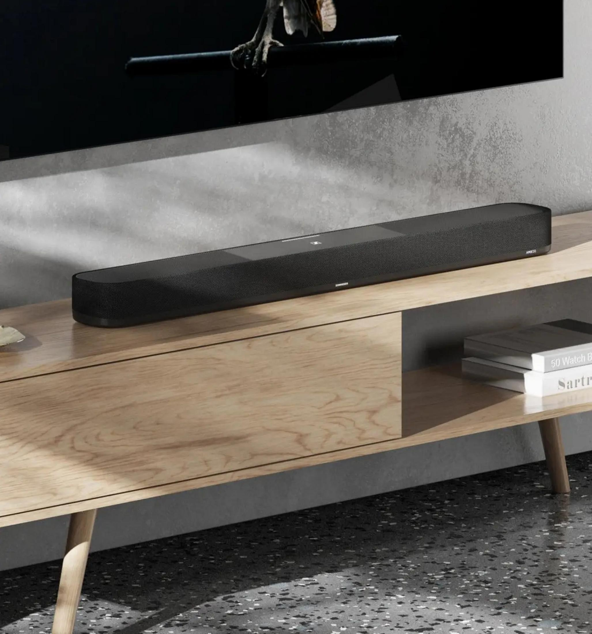Ein schlanker schwarzer Soundbar steht auf einem Holzmöbel vor einem Fernseher.