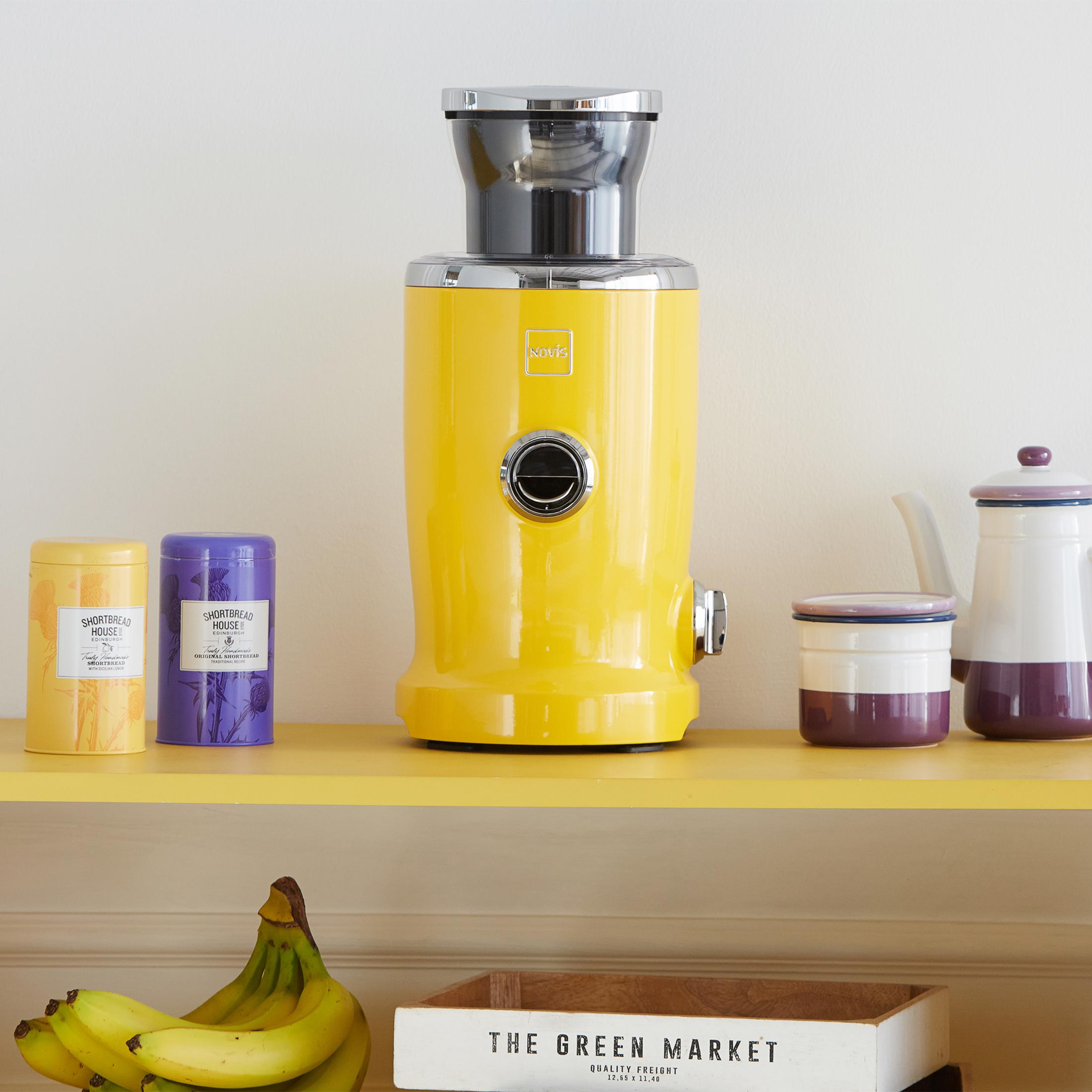 Ein gelber Entsafter steht auf einem Regal mit Dosen und einer Teekanne. Unten sind Bananen.