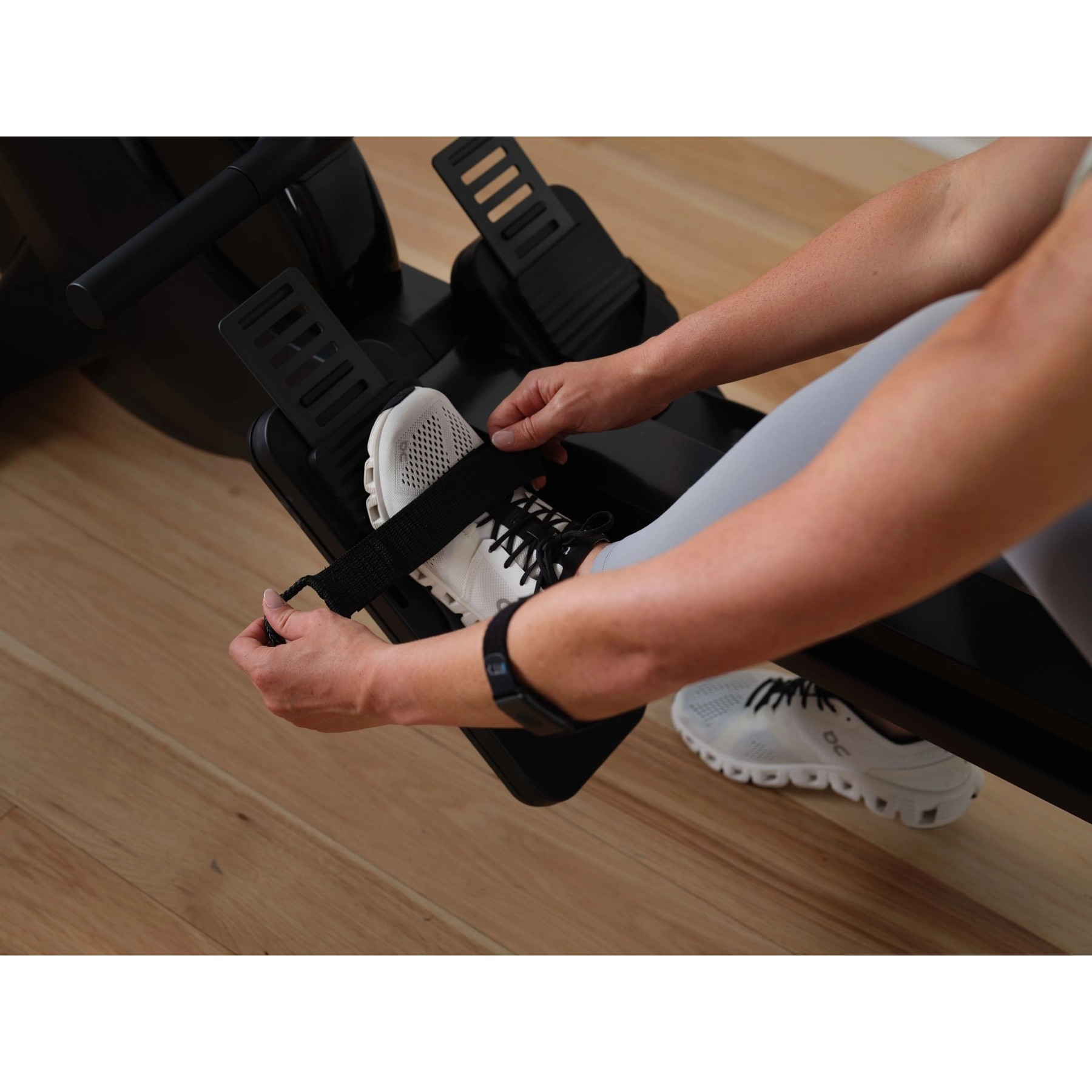 Person befestigt Fußgurt an einem Ergometer. Weiße Schuhe, schwarzer Gurt.