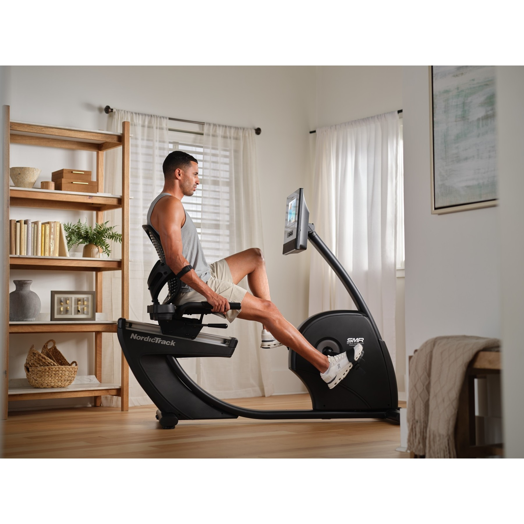 Ein Mann trainiert auf einem schwarzen Heimtrainer in einem hellen Raum mit einem Regal.