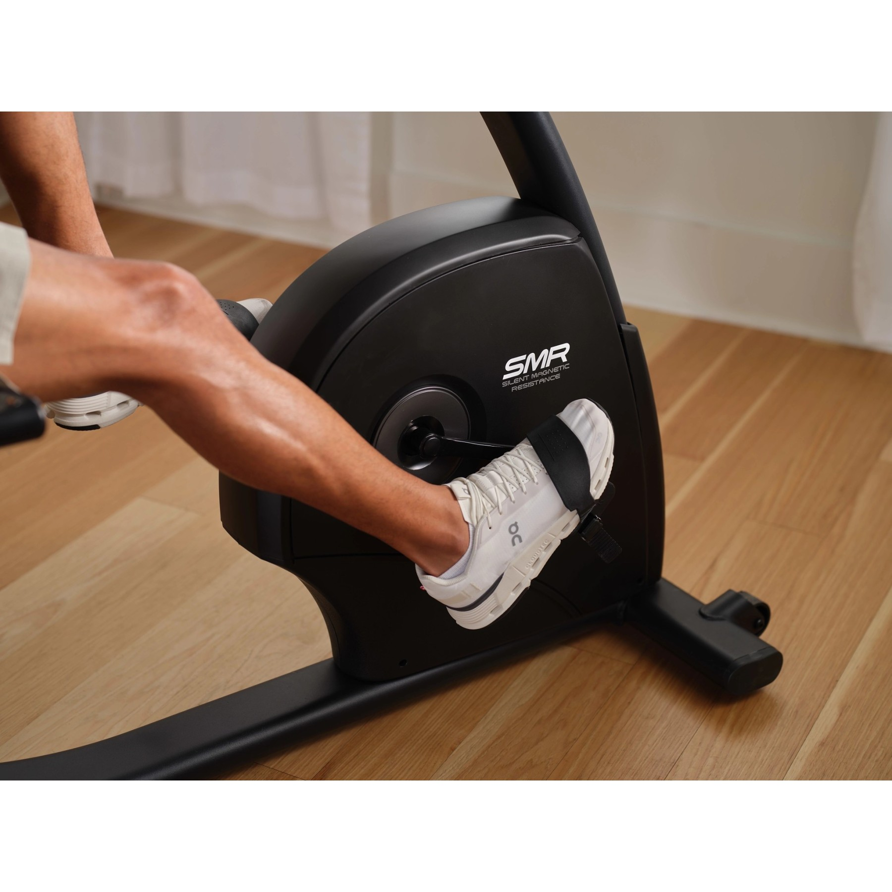 Ein Mann tritt auf einem schwarzen Heimtrainer mit weißen Turnschuhen auf einem Holzboden.