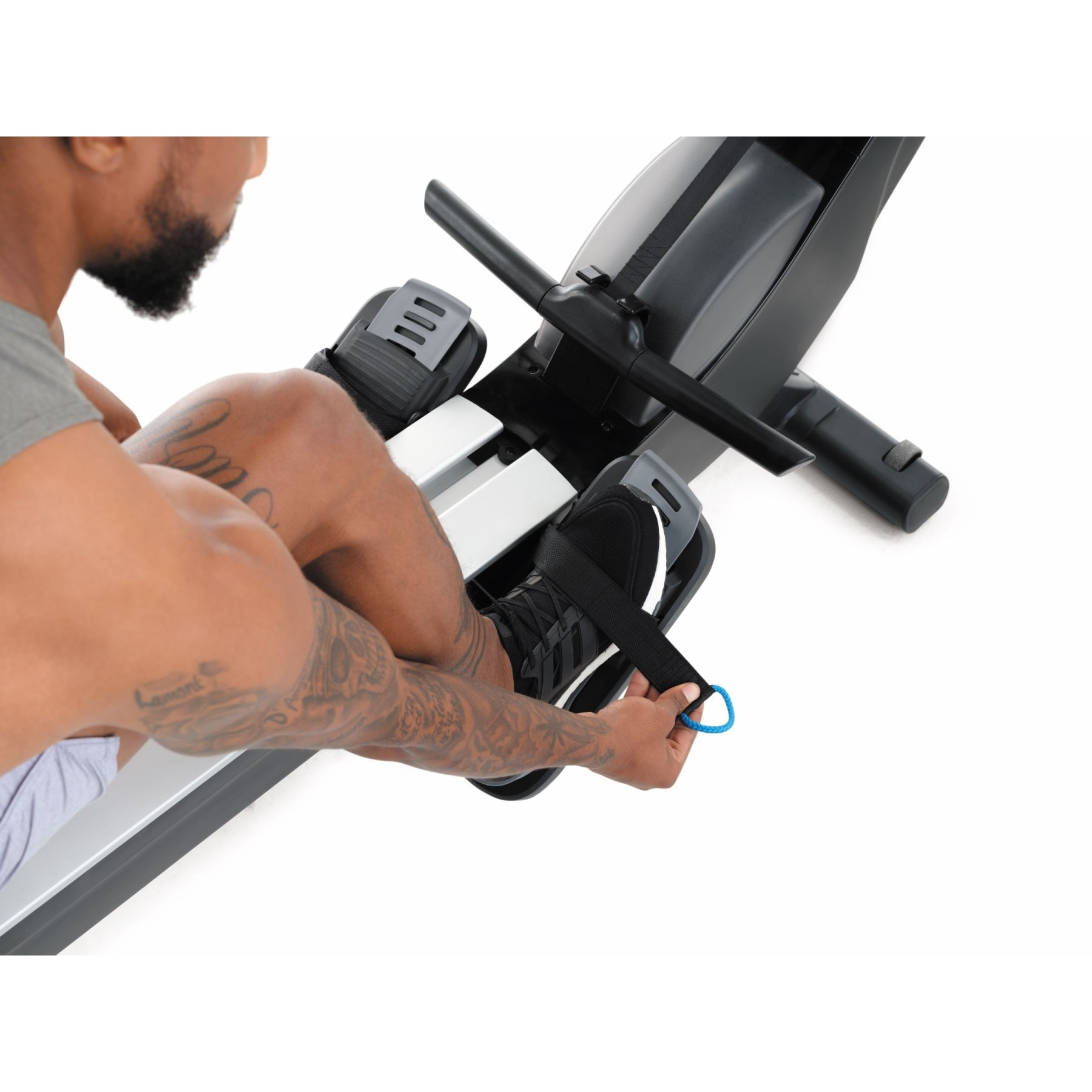 Mann mit Tattoos an Beinen und Arm benutzt Rudergerät, sichert Fußriemen.