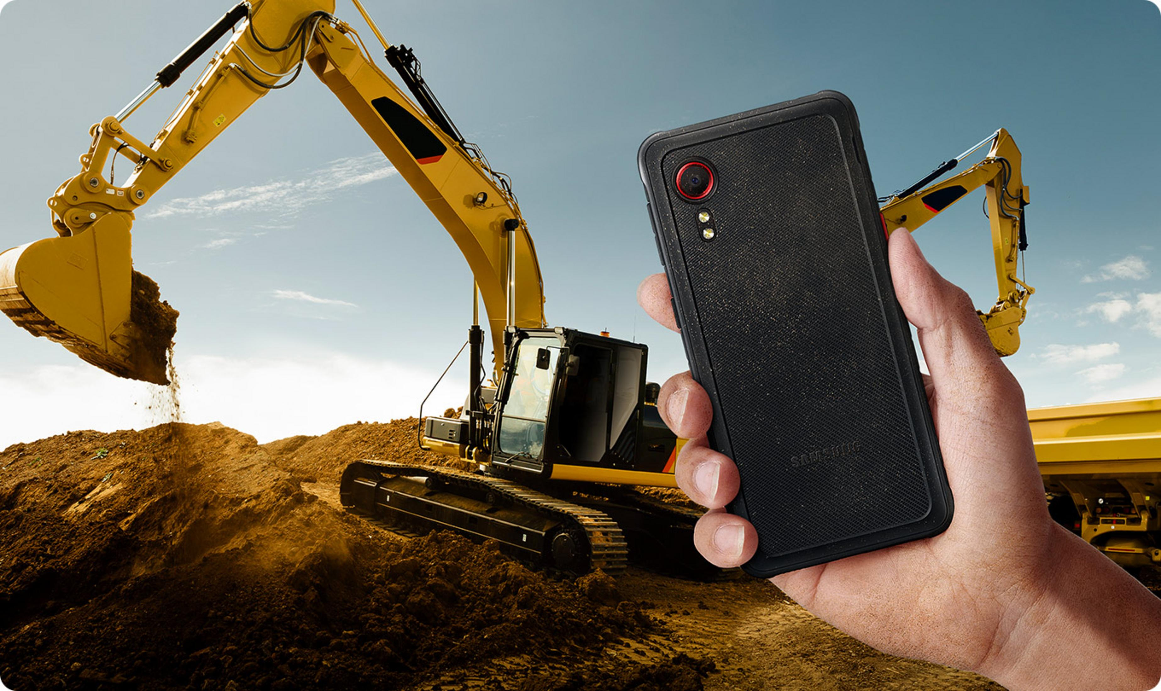 Una mano sostiene un teléfono negro frente a excavadoras y un camión volquete amarillo.