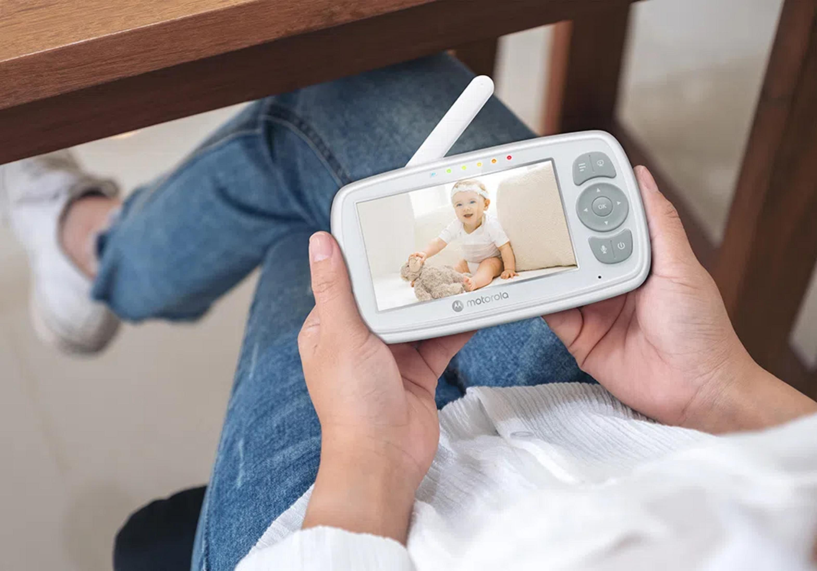 Baby auf Monitorbildschirm spielt mit Teddybär, gehalten von Person in Jeans.