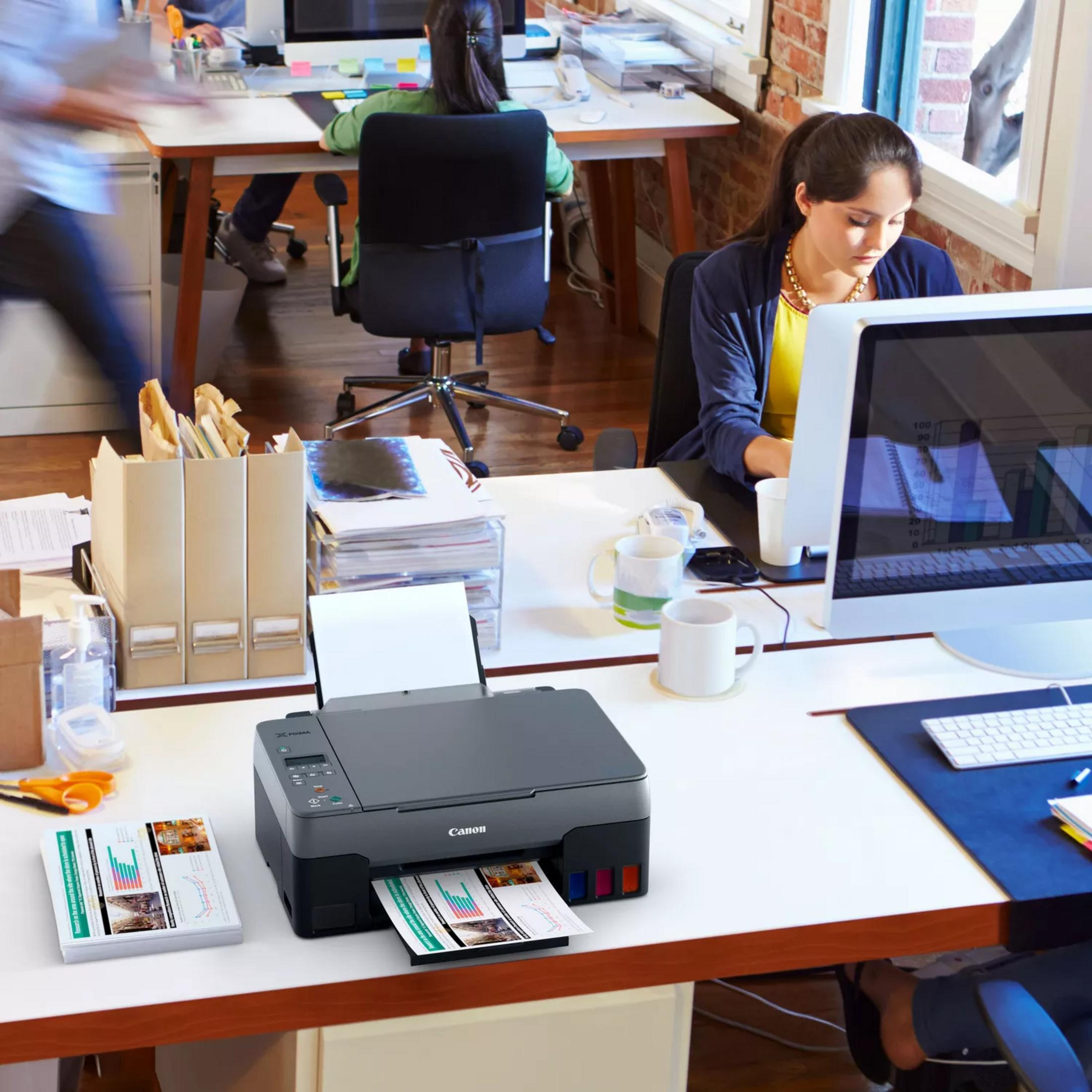 Scena d'ufficio con stampanti, computer e persone al lavoro.