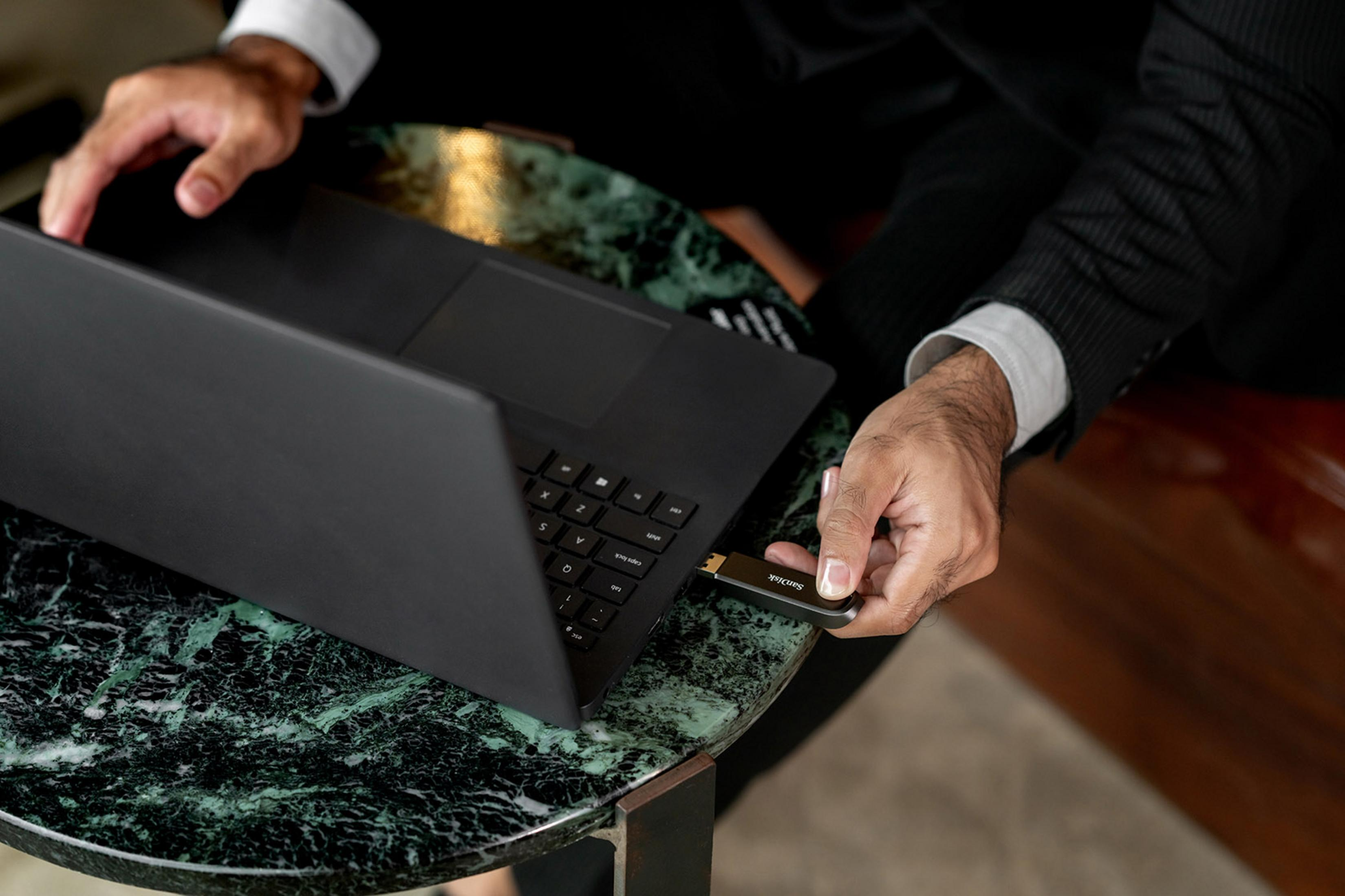Uomo inserisce chiavetta SanDisk in laptop nero su tavolo marmo.