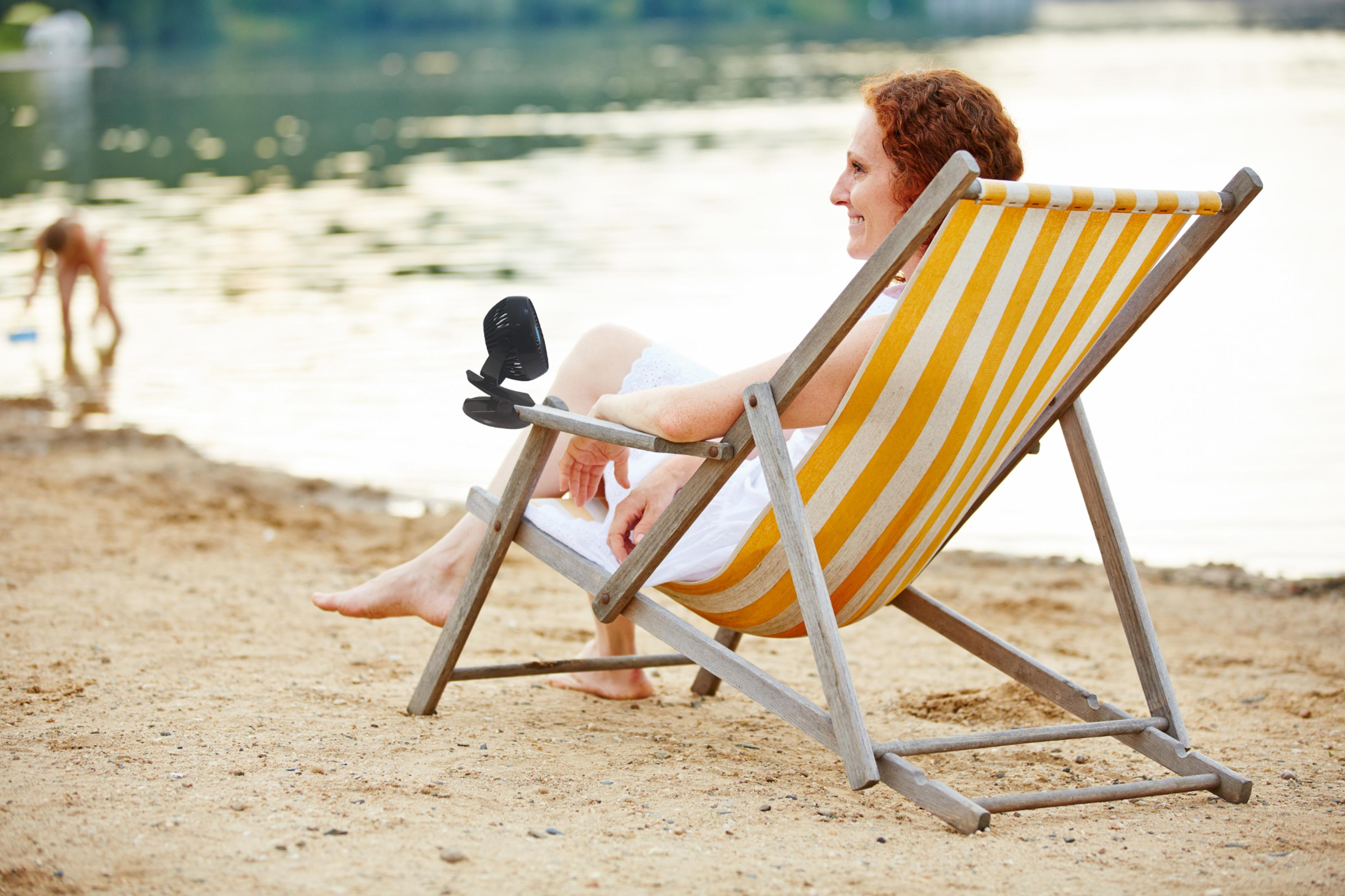 Frau sitzt im Liegestuhl am Strand, trinkt aus einer Tasse, im Hintergrund ein Kind.