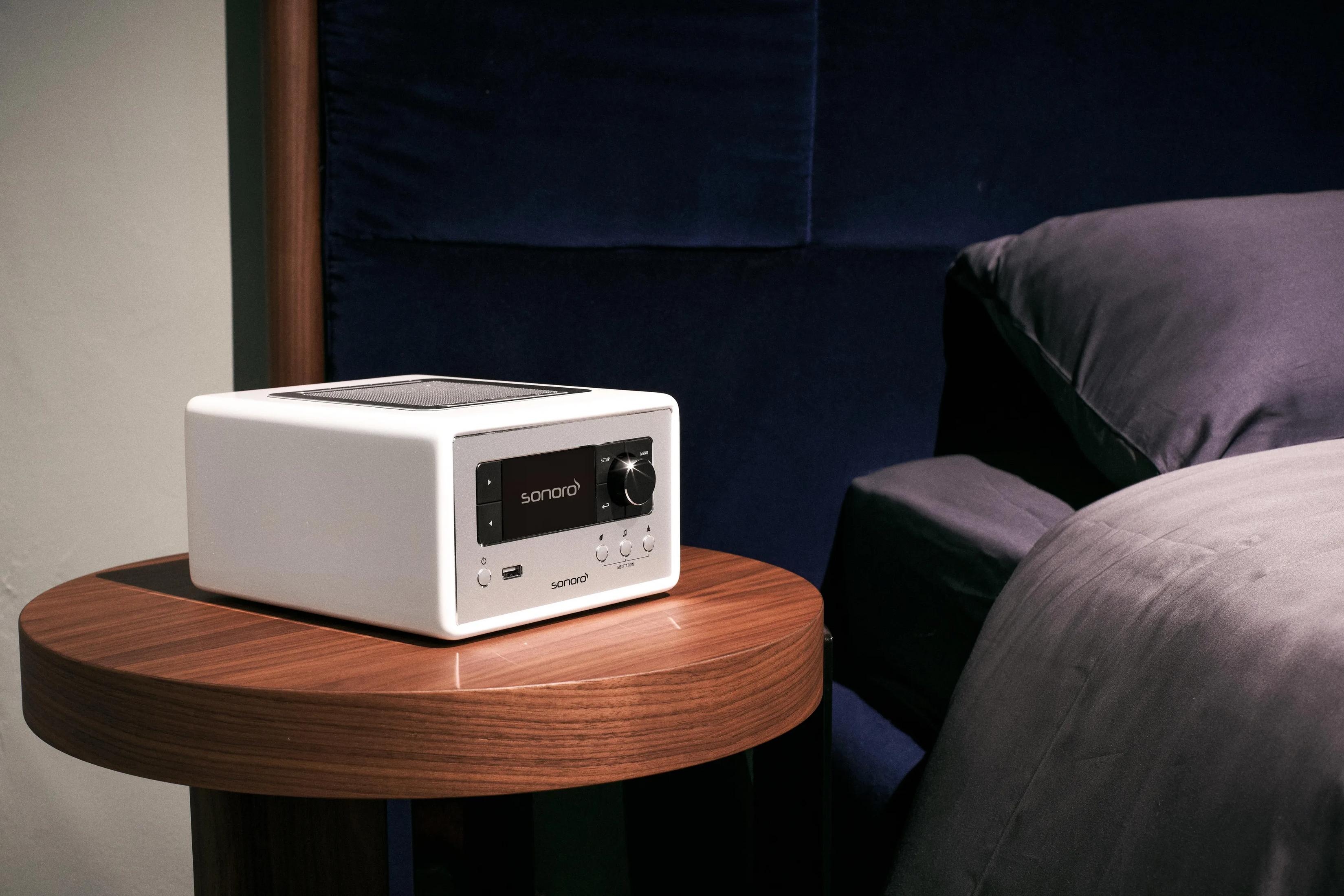 A white radio sits on a round wooden table next to a bed.