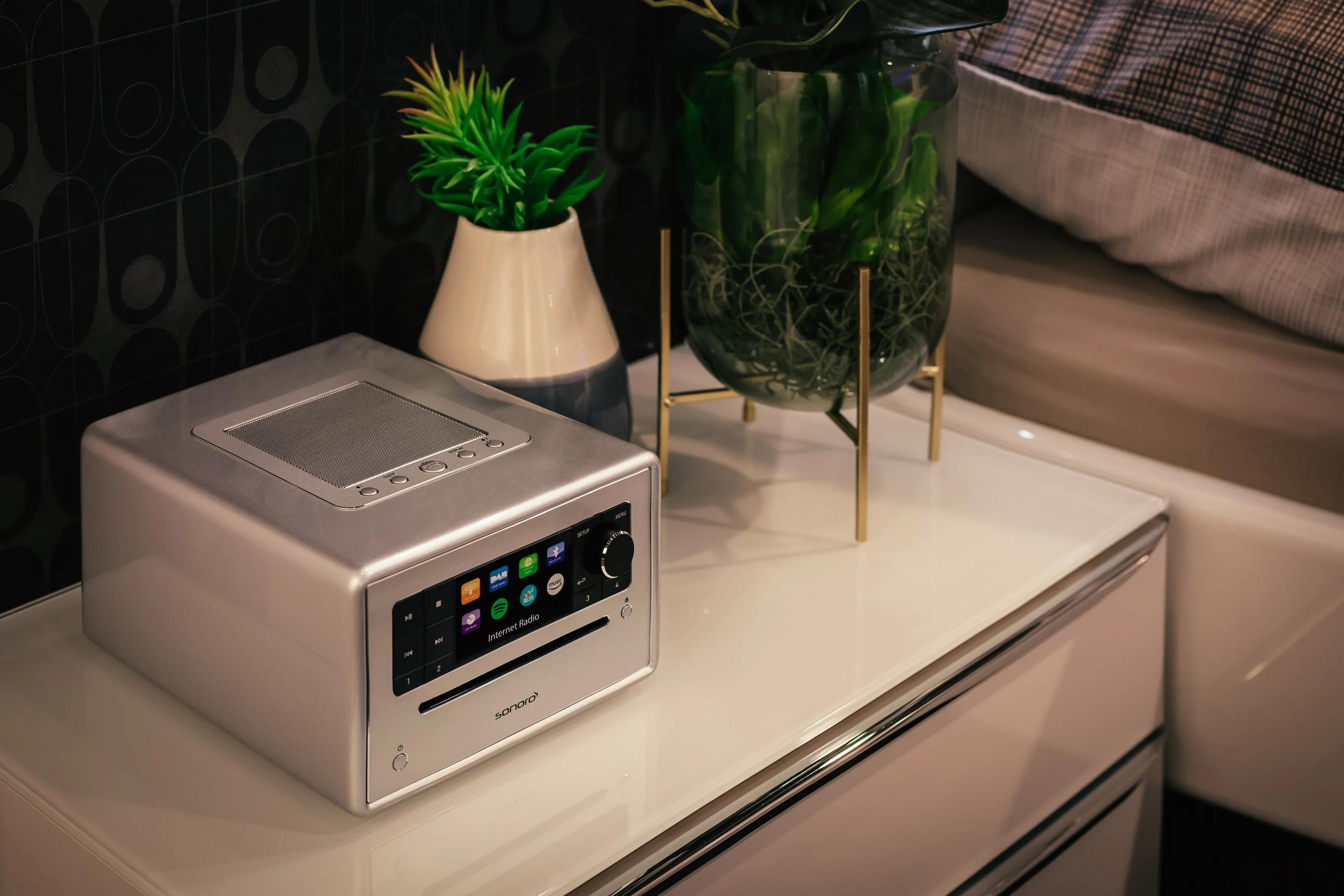 Een zilveren radio met touchscreen staat op een nachtkastje naast planten.