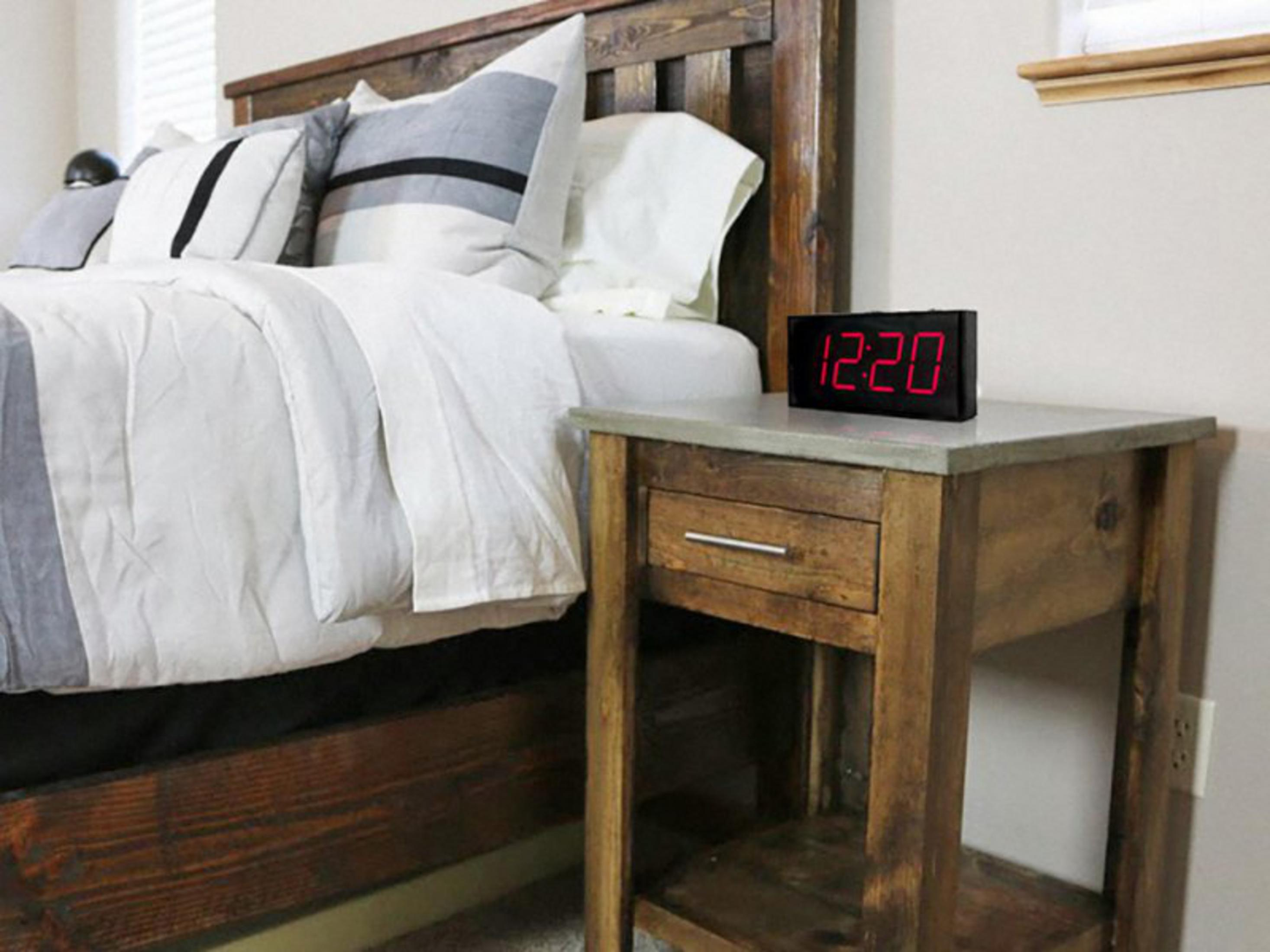 Un lit en bois avec des oreillers, à côté une table de nuit avec une horloge numérique (12:20).