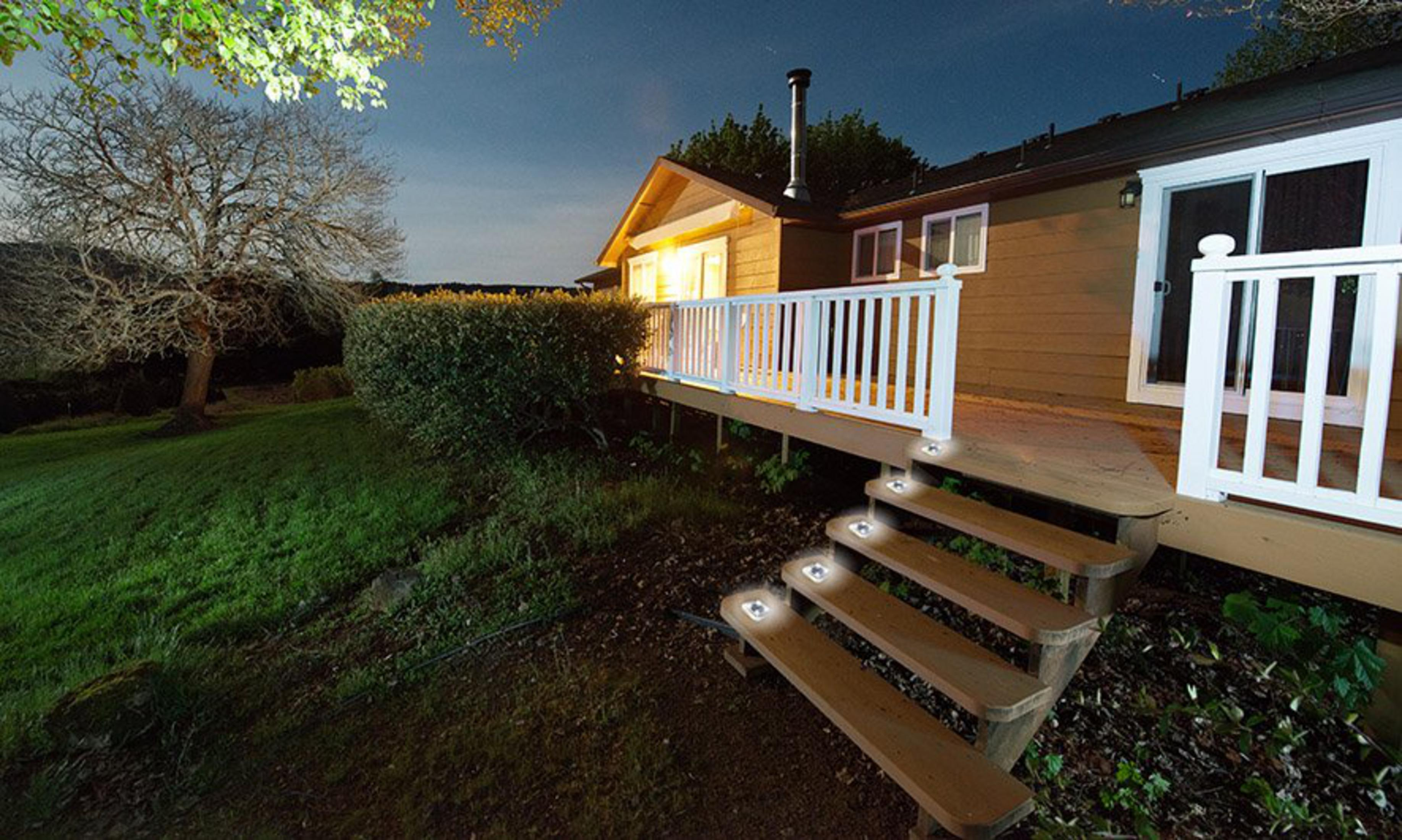 Une terrasse en bois avec des marches éclairées mène à une maison la nuit. Un arbre et une pelouse sont visibles.