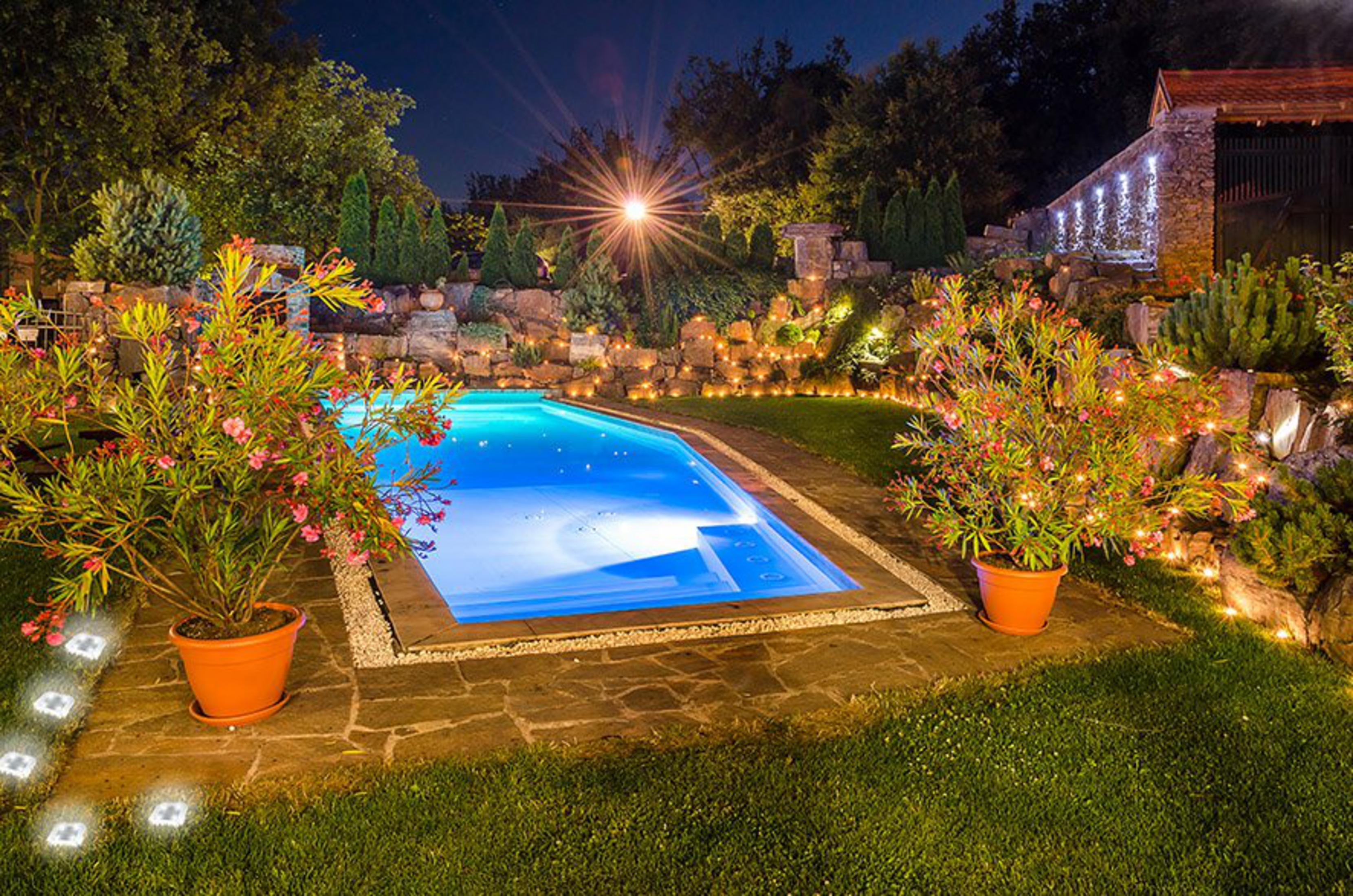 Une piscine bleue illuminée la nuit, avec des plantes en pot et des murs en pierre éclairés.