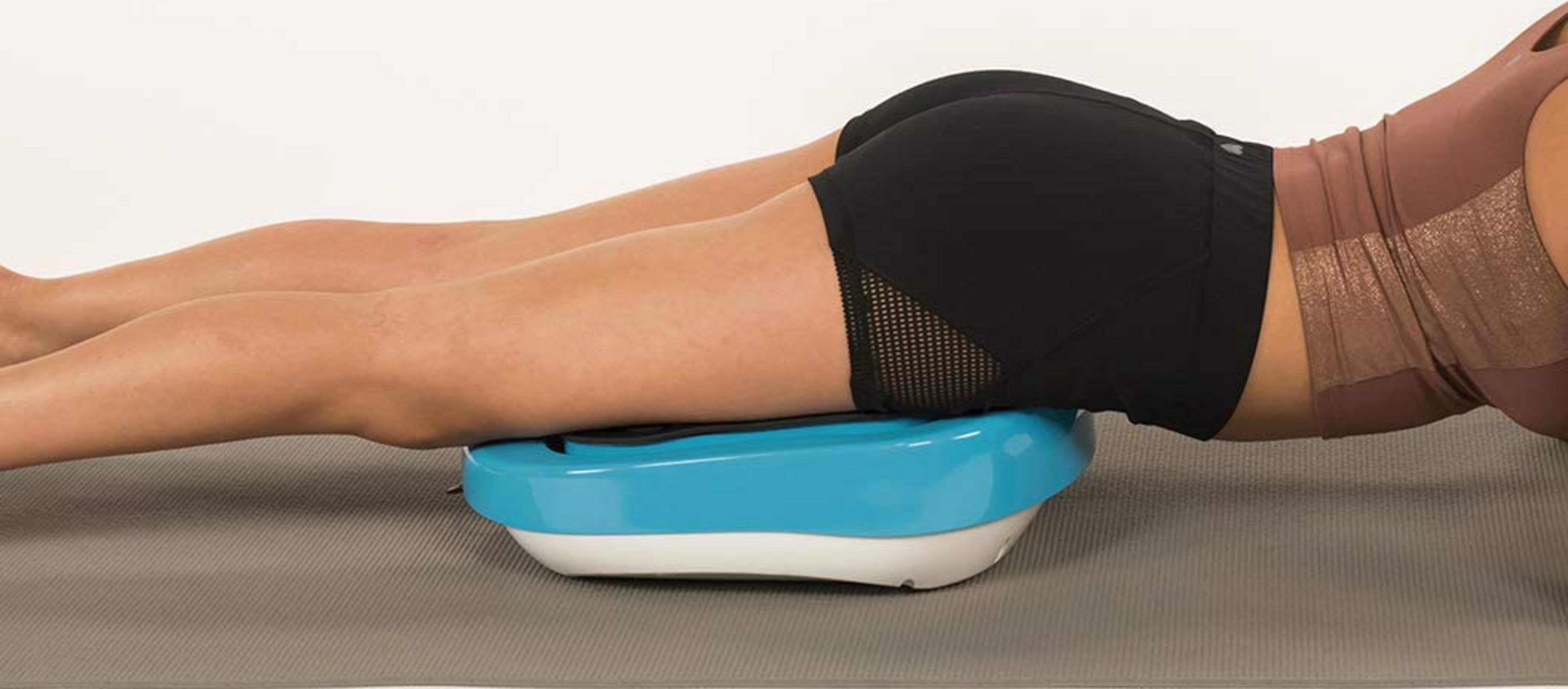 Eine Frau liegt auf einem Vibrationsgerät in Schwarz und Blau auf einer Matte.