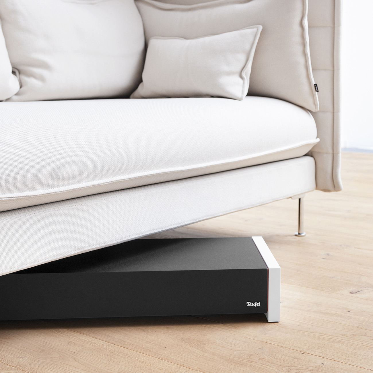 Ein schwarzer Subwoofer mit weißem Rand unter einem beigen Sofa auf einem Holzboden.