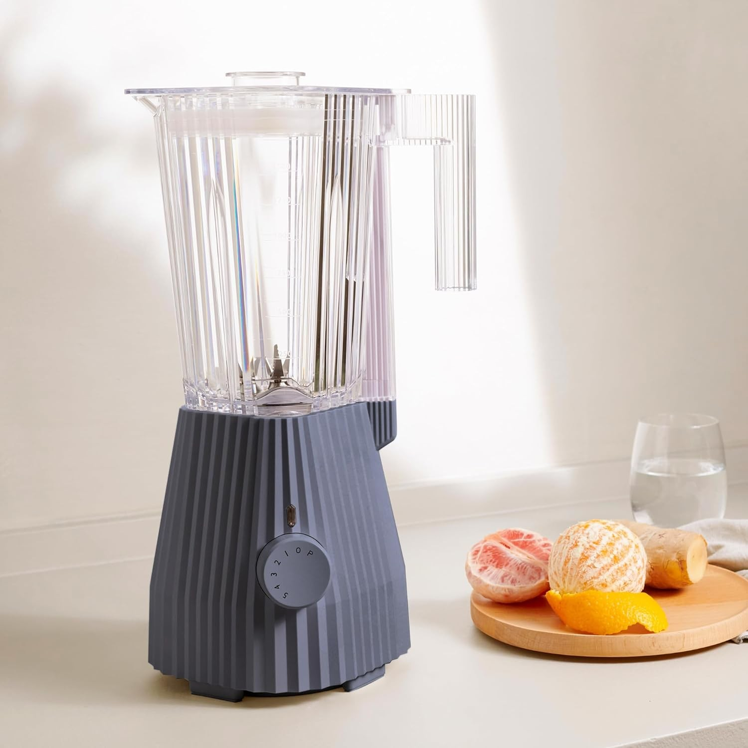 Un frullatore grigio accanto a frutta su un piatto di legno. Un bicchiere d'acqua sullo sfondo.