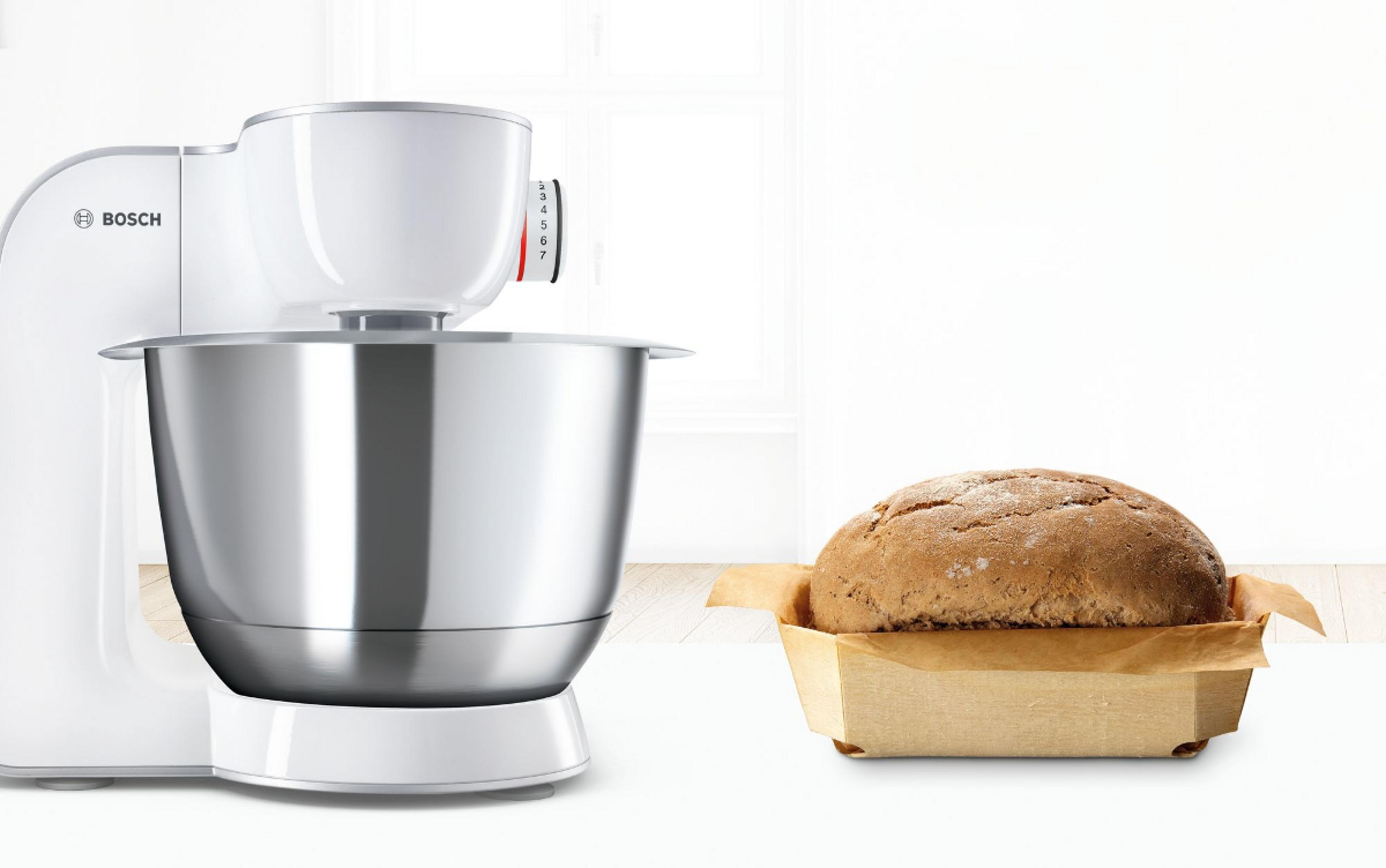 Eine weiße Bosch Küchenmaschine steht neben einem Laib Brot in einer Backform.