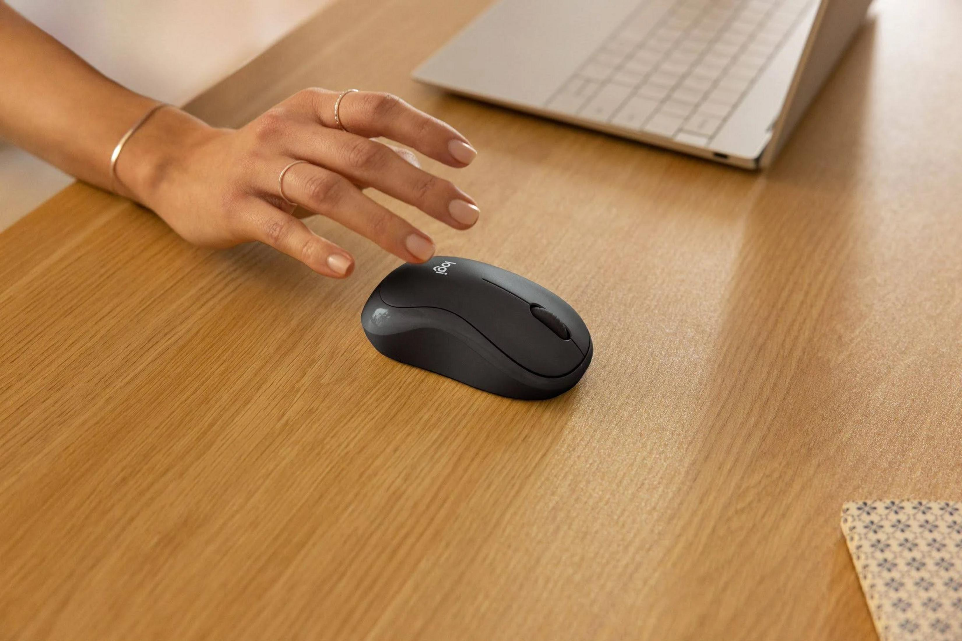 Dłoń z pierścionkami używa czarnej myszy Logitech obok laptopa.
