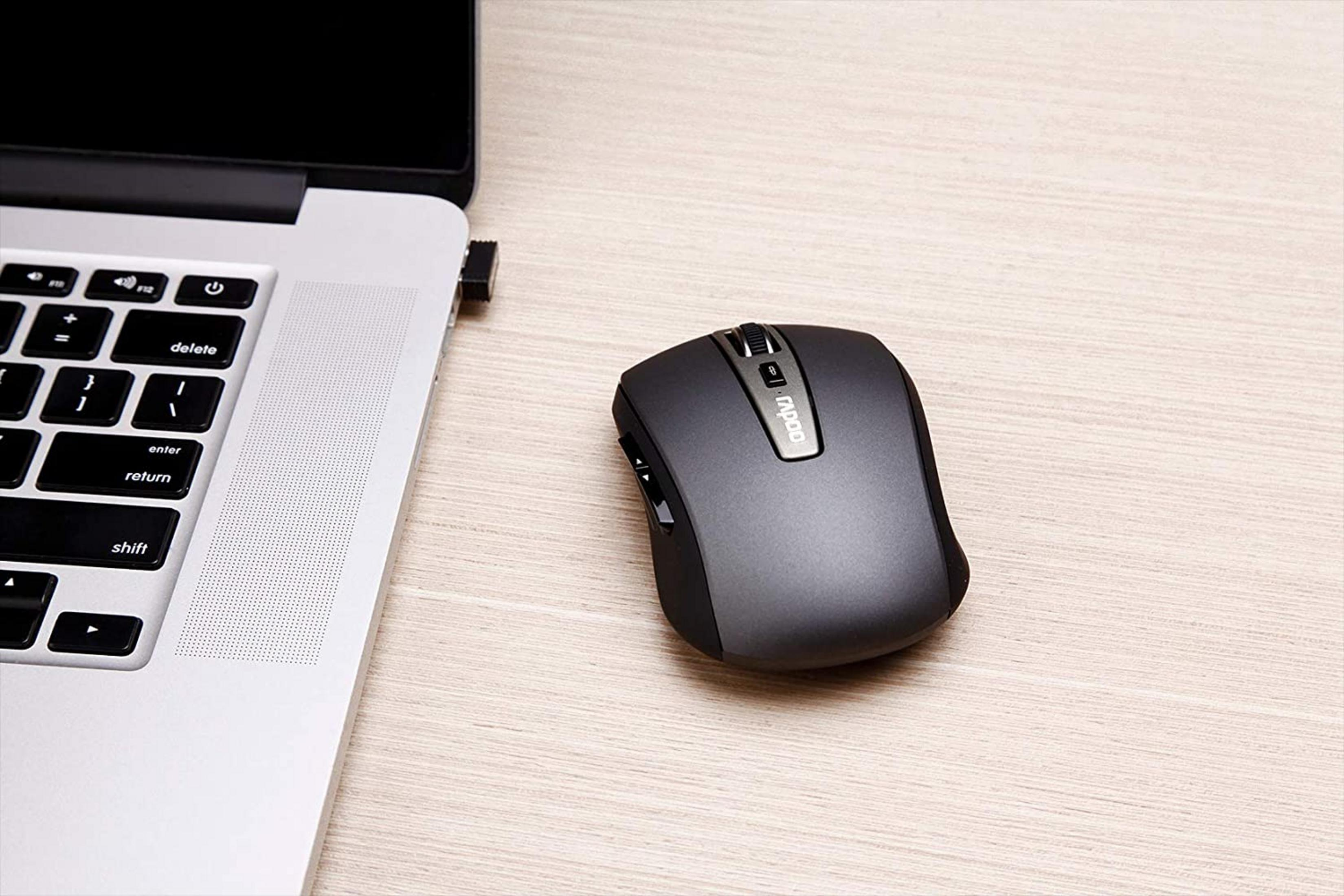 Een donkere muis naast een zilveren laptop op een houten bureau.