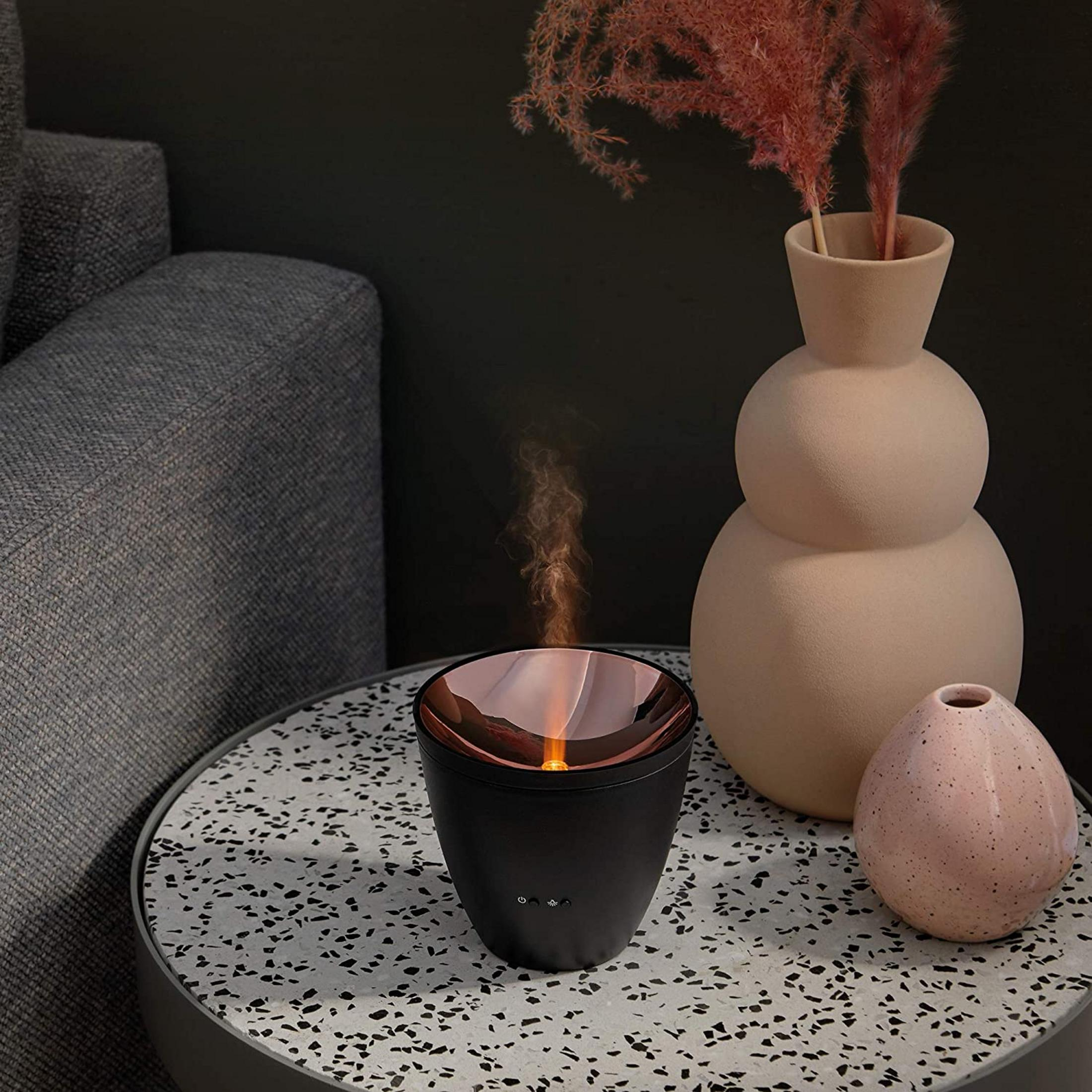 Ein schwarzer Diffusor mit Flammenoptik steht auf einem Terrazzo-Tisch neben Vasen.