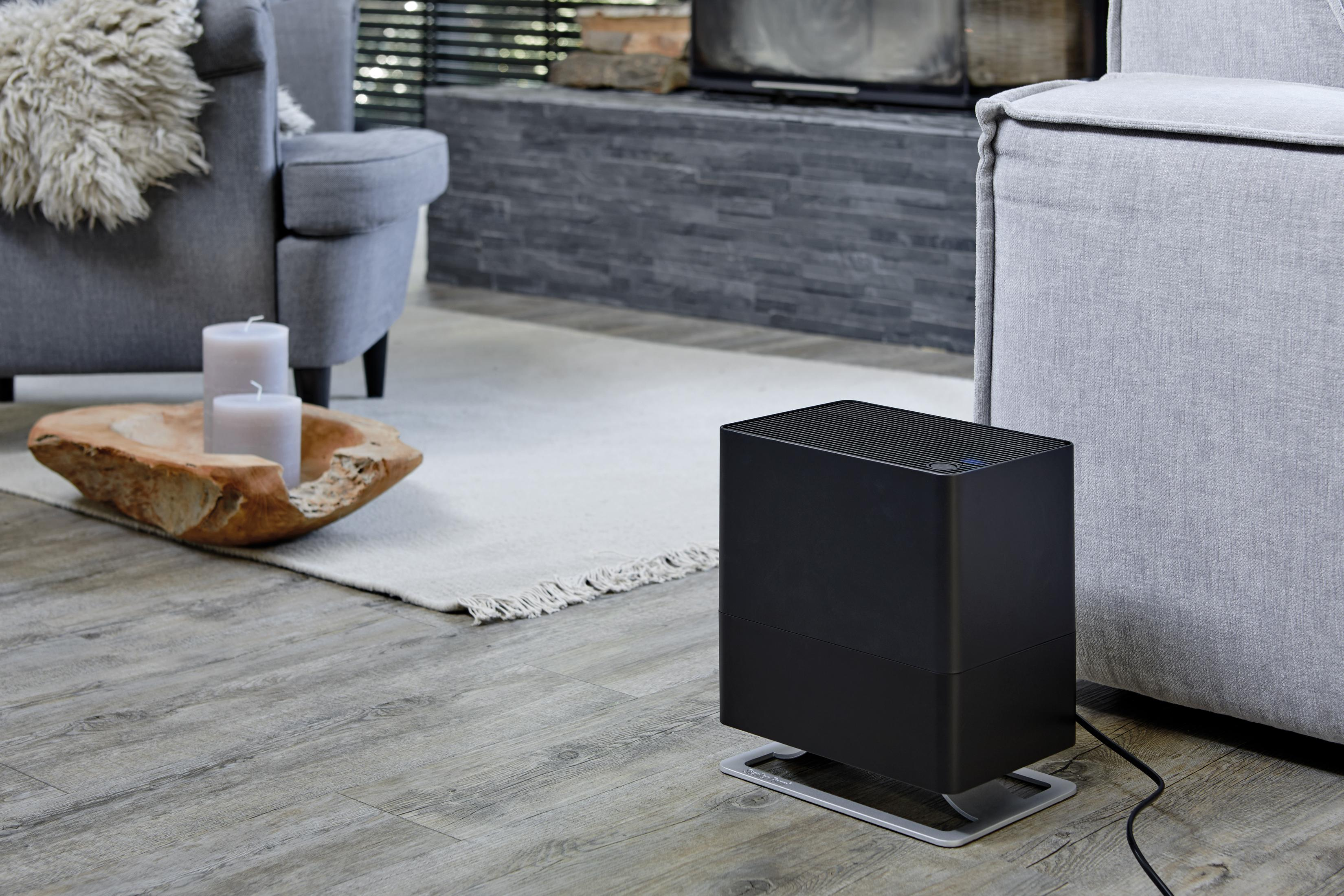 A black humidifier sits on a wooden floor next to a grey sofa.