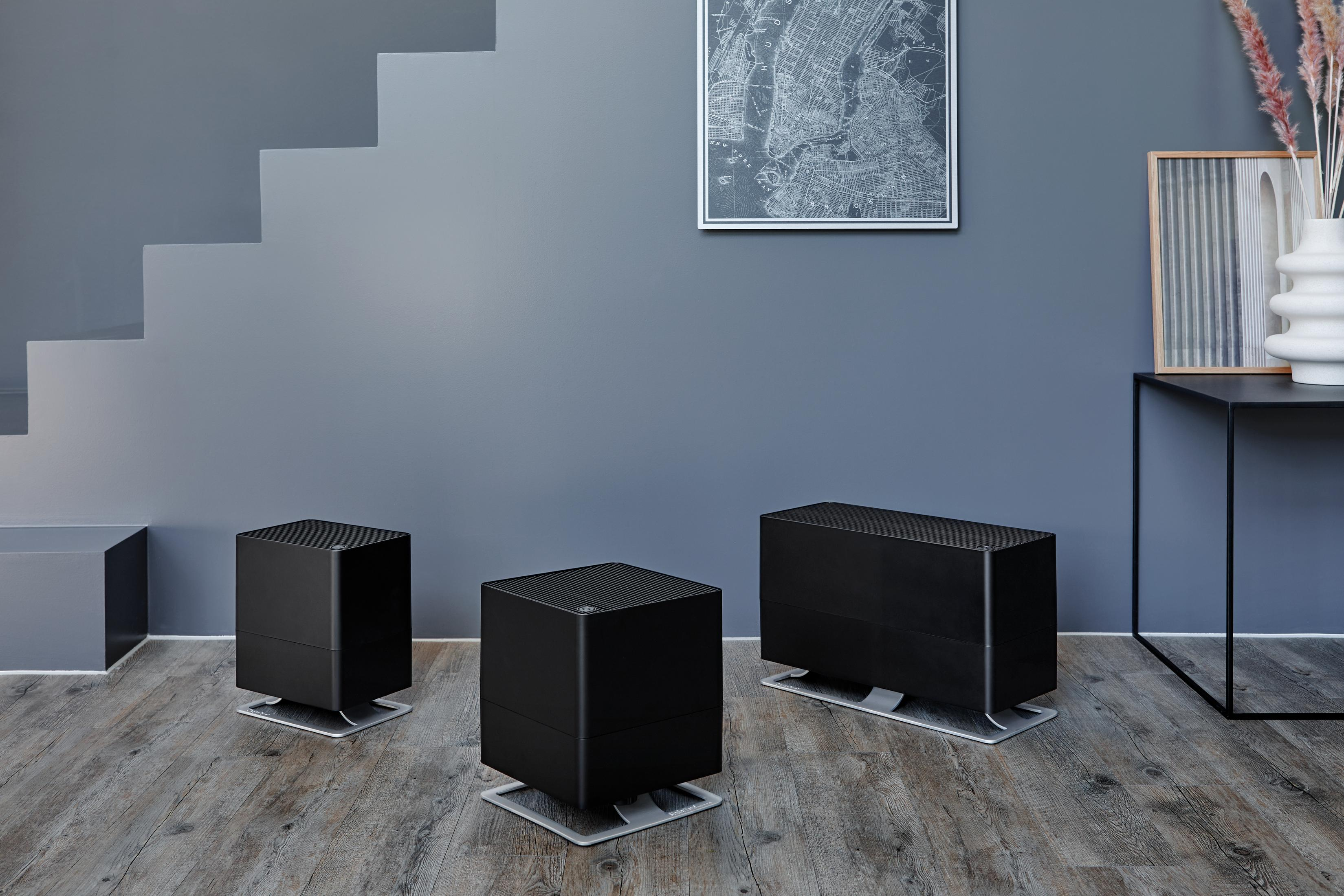 Three black humidifiers on a wooden floor, with stairs and wall art.