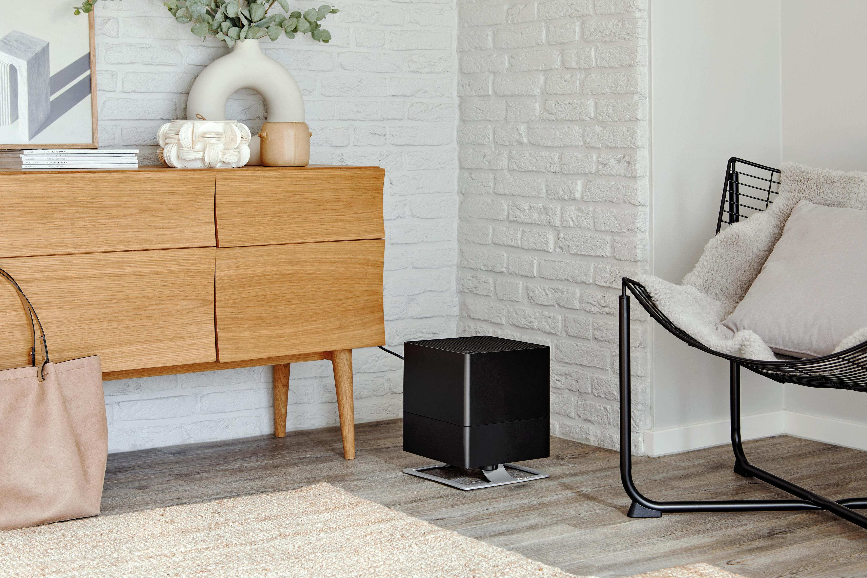 A black humidifier sits on a wood floor next to a wooden dresser and a black chair.