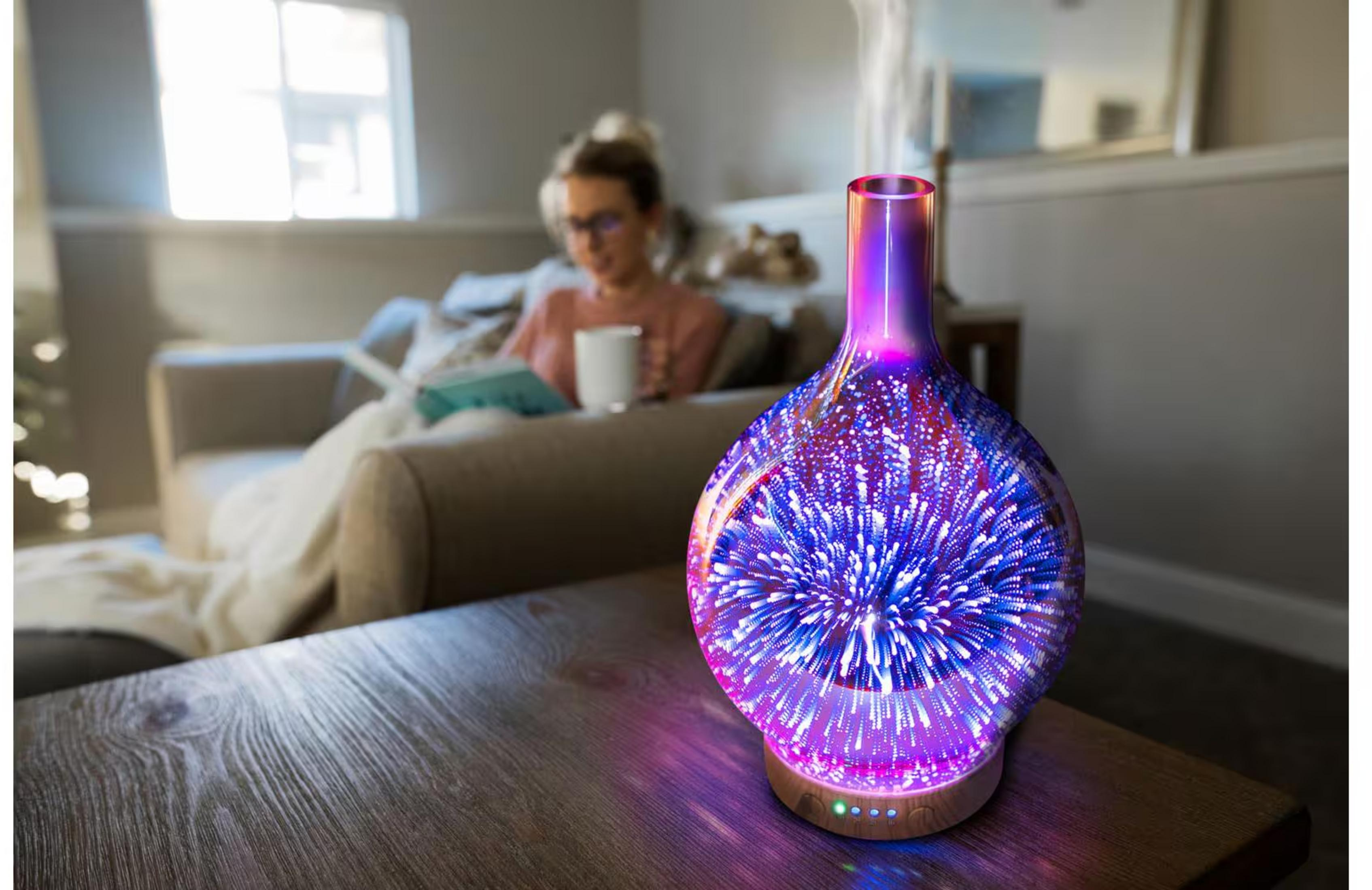 Ein aromatischer Diffusor mit Sternenmuster leuchtet lila und blau. Eine Frau liest im Hintergrund.