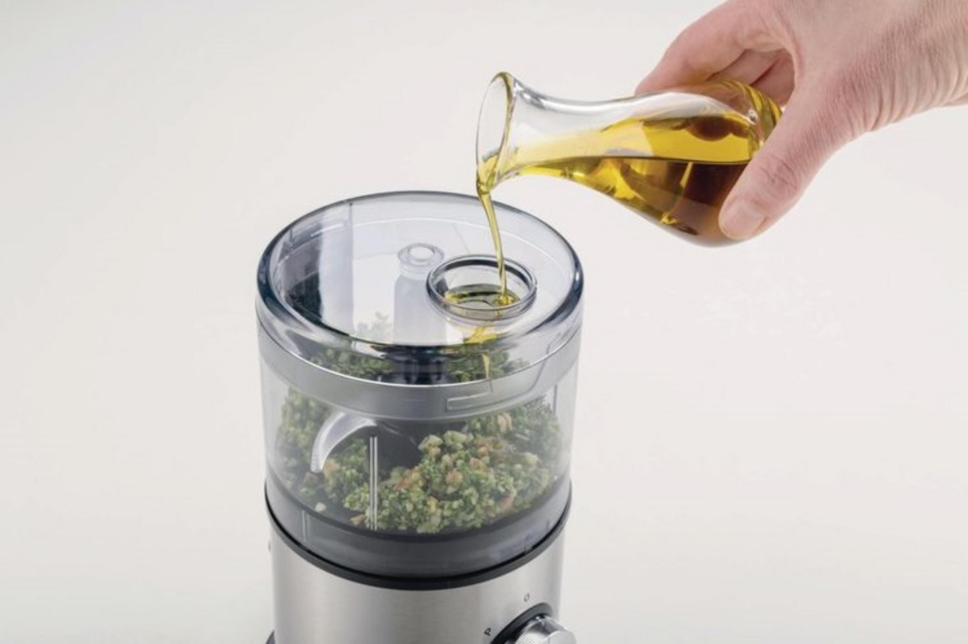 L'olio d'oliva viene versato in un robot da cucina con ingredienti verdi.