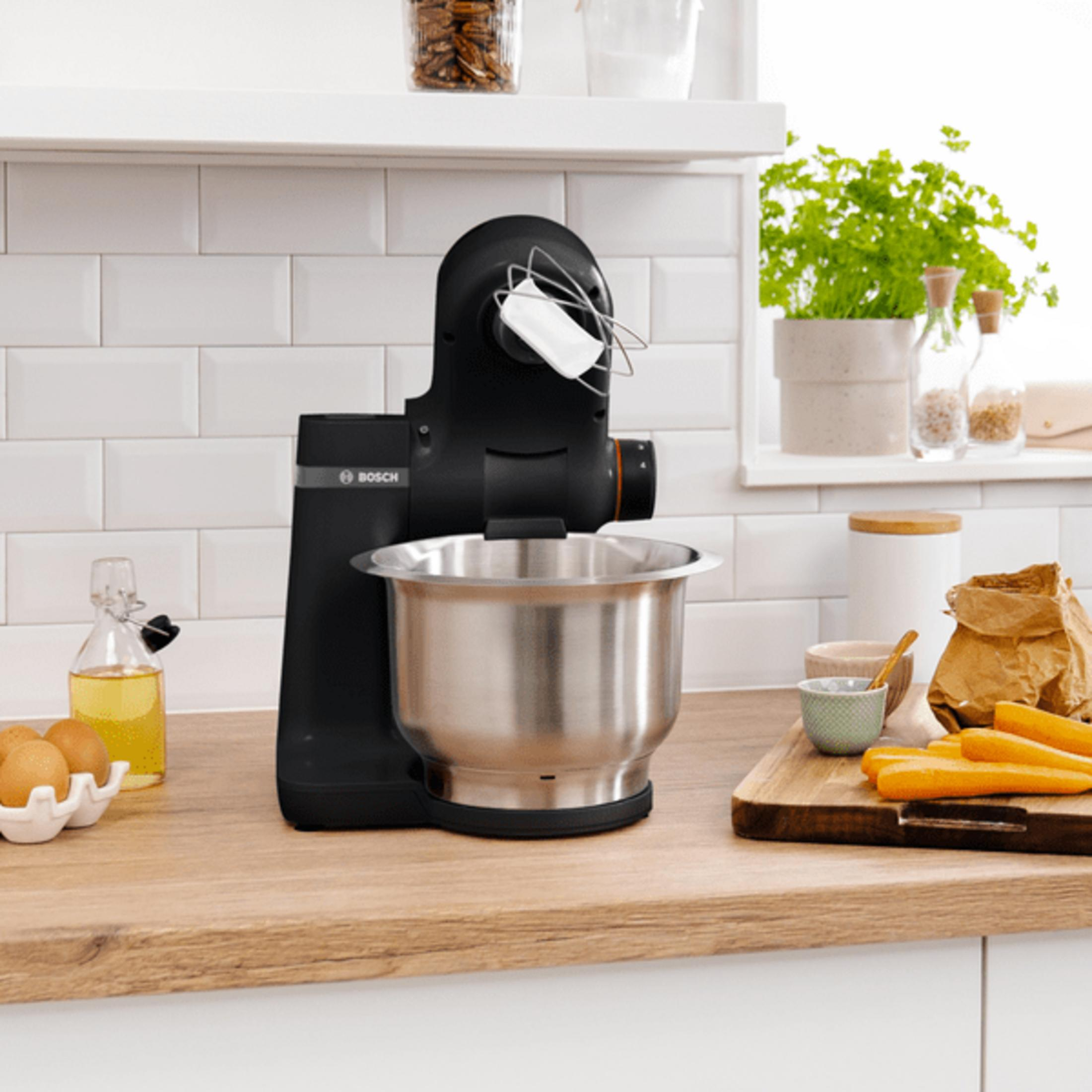 Schwarzer Küchenmixer mit Edelstahlschüssel, Eiern, Öl und Karotten.