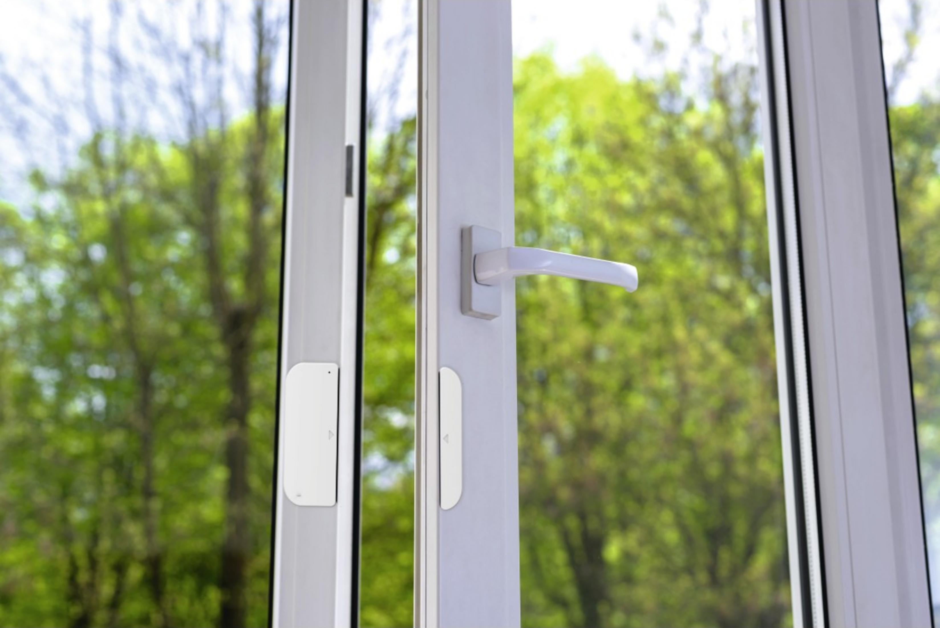 Un tirador de ventana blanco y sensores en un marco de ventana blanco con árboles detrás.
