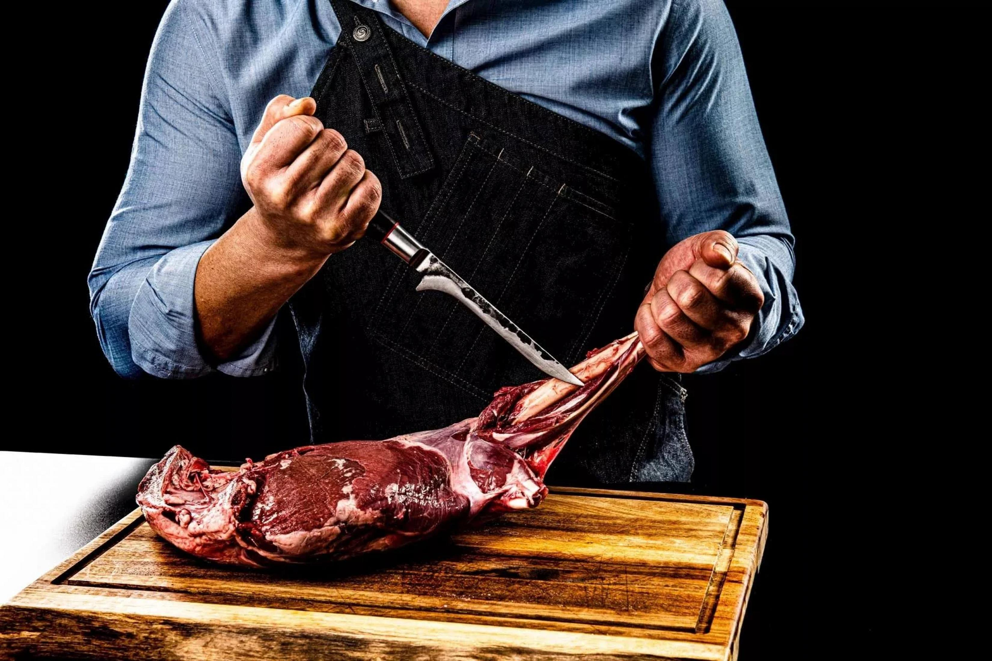 Un carnicero con camisa azul y delantal negro está cortando carne cruda en una tabla de cortar.