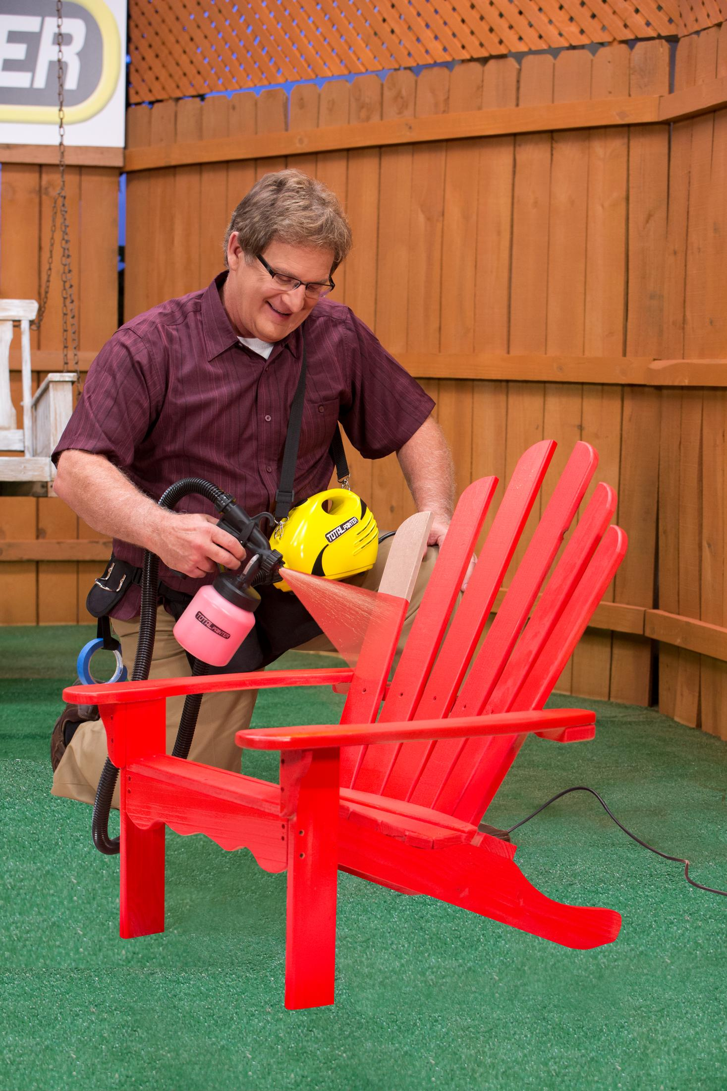 Mann sprüht roten Adirondack-Stuhl mit Sprühgerät in einem Garten.