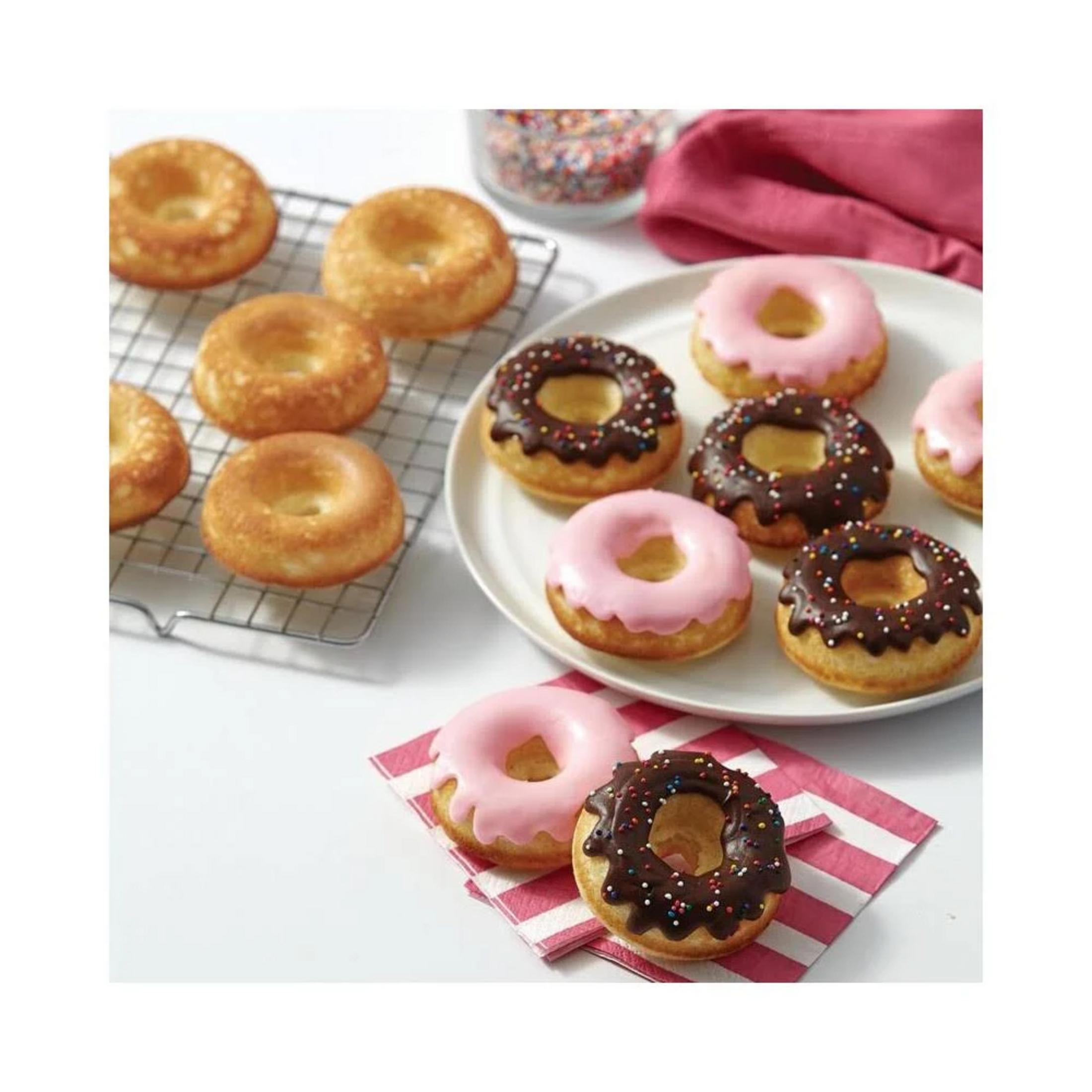 Drei rosa und drei braune Donuts mit Streuseln auf einem Teller. Mehr Donuts auf einem Rost.
