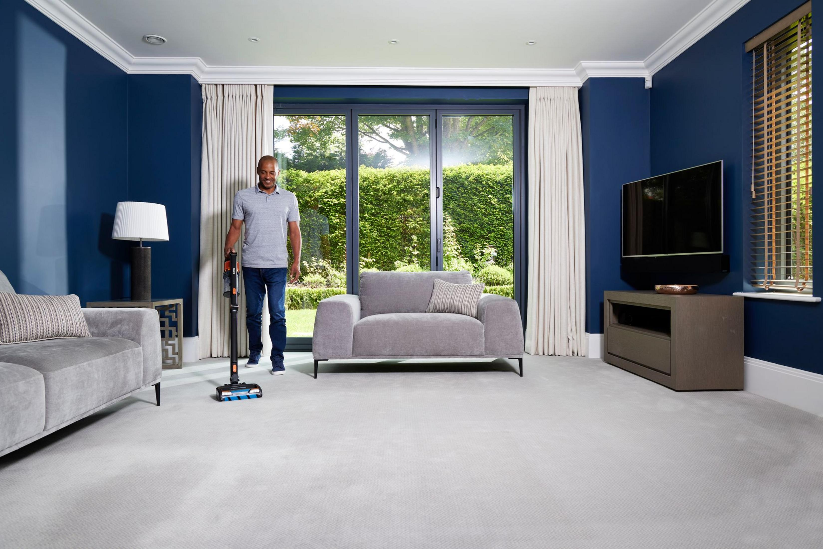 Hombre aspira alfombra en sala azul con sofá y TV.