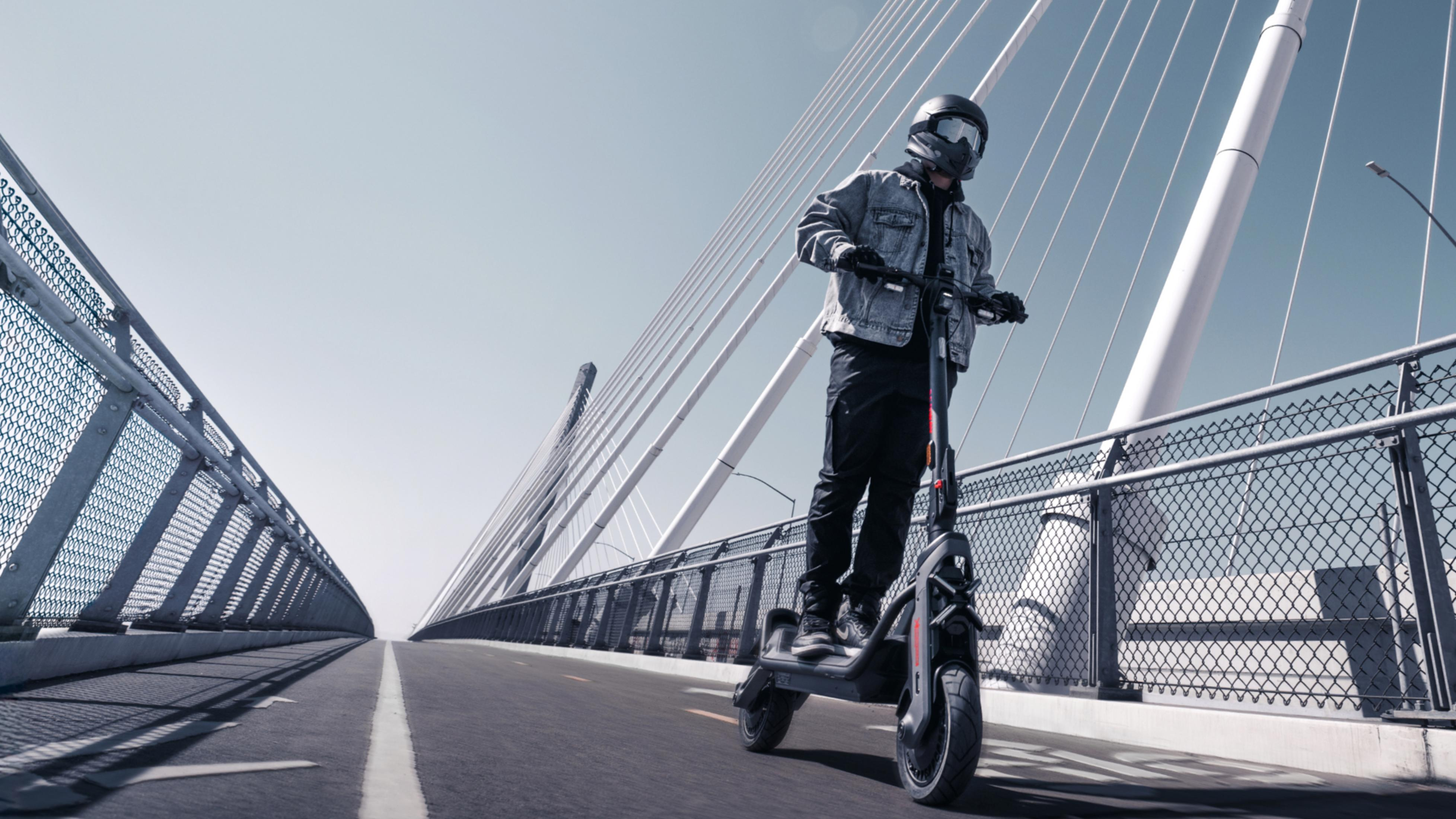 Mann in Jeansjacke fährt E-Scooter auf Brücke mit Kabeln vor Himmel.