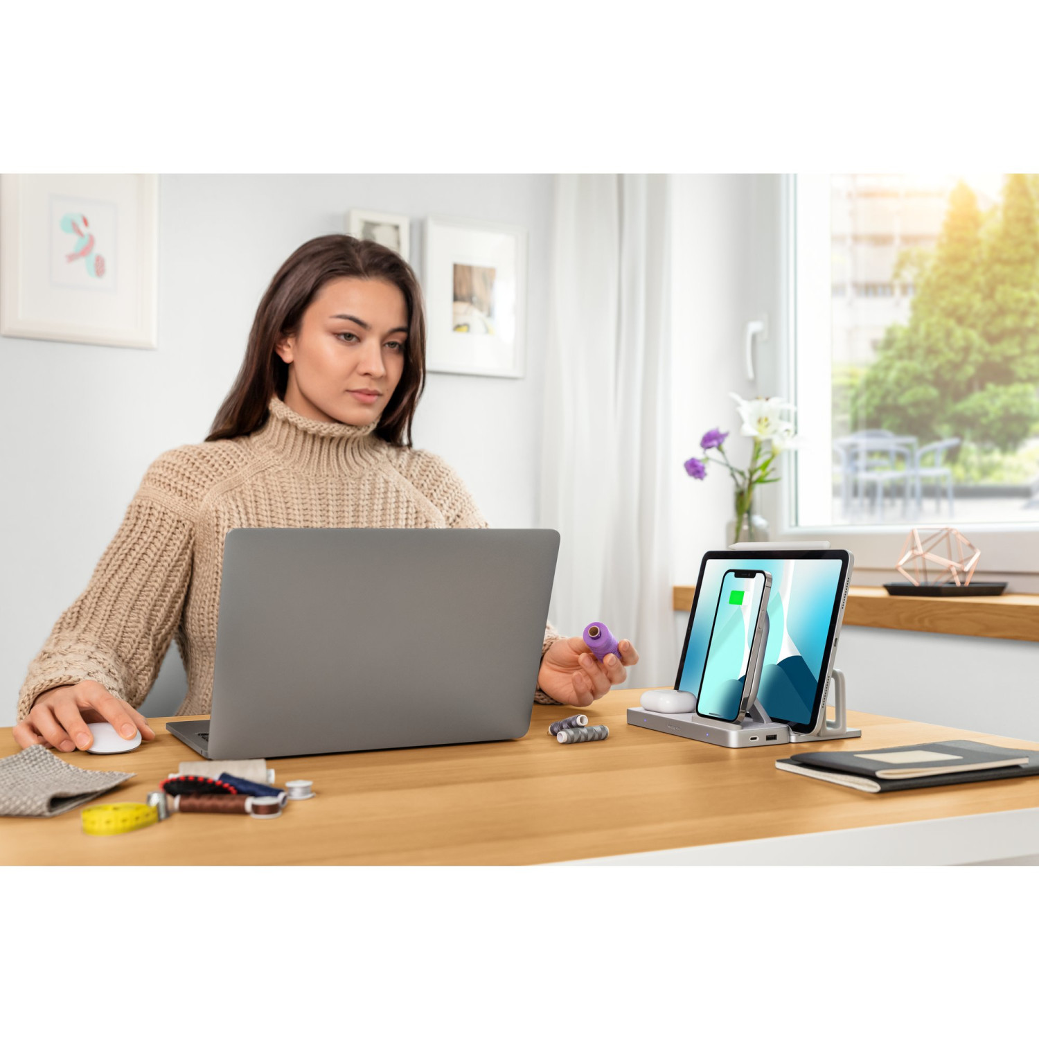 Frau am Schreibtisch mit Laptop, Handy wird auf Ständer geladen. Nähsachen in der Nähe.