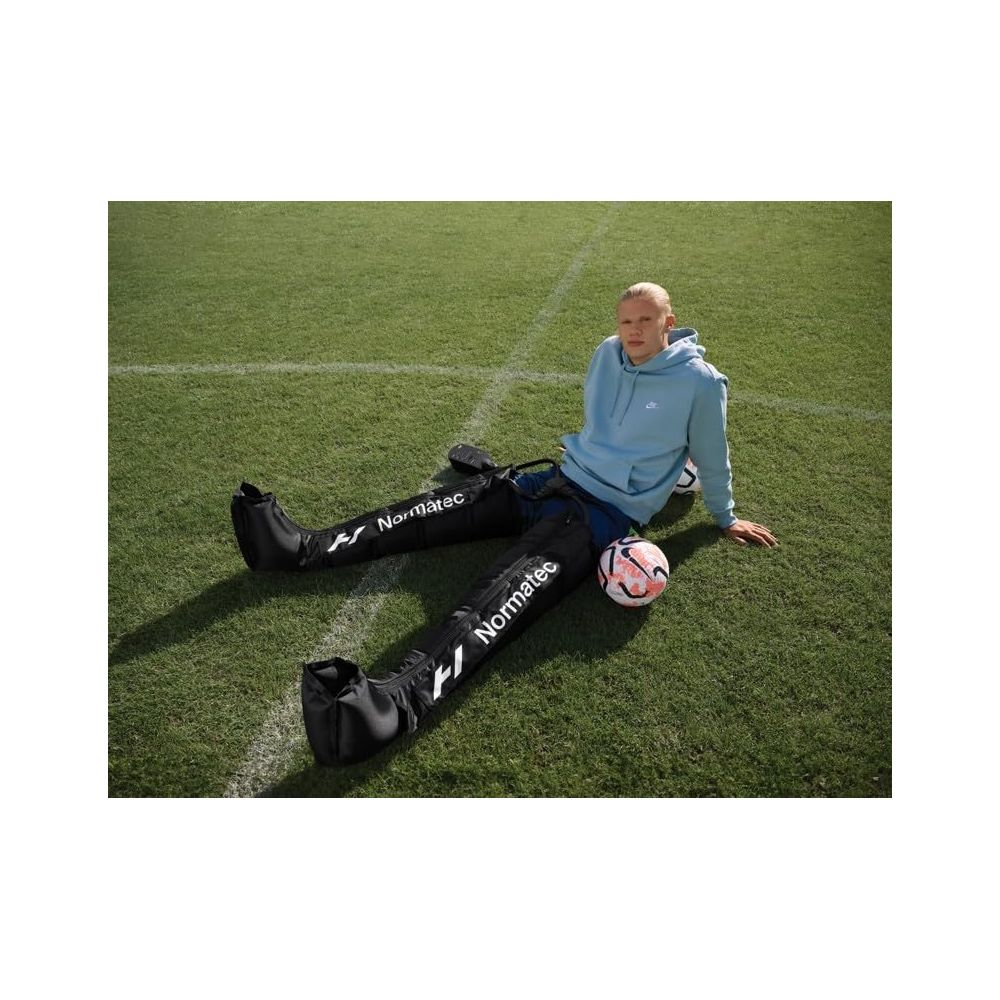 Ein Mann sitzt auf einem Fußballfeld mit komprimierenden Beinstulpen und einem Ball.