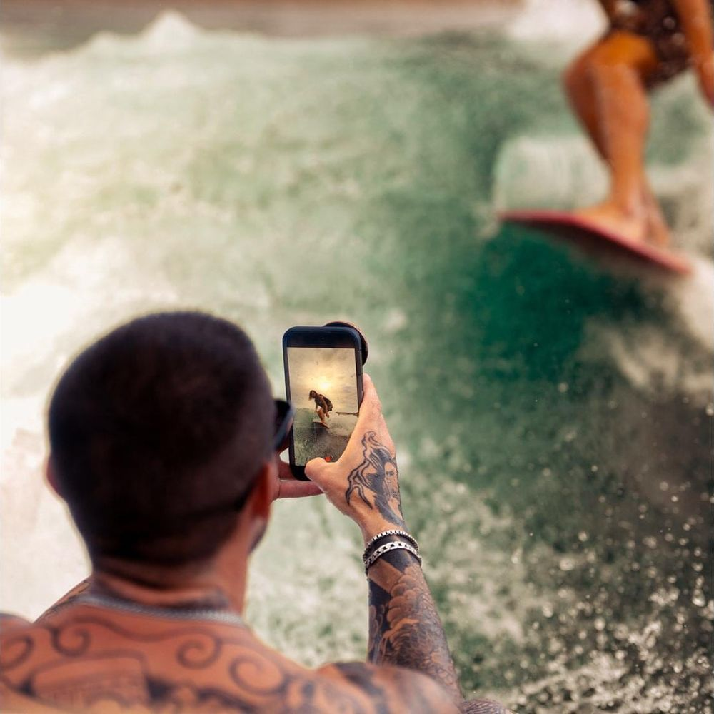 Ein Mann mit Tattoos fotografiert einen Surfer auf dem Handy. Wasser spritzt.