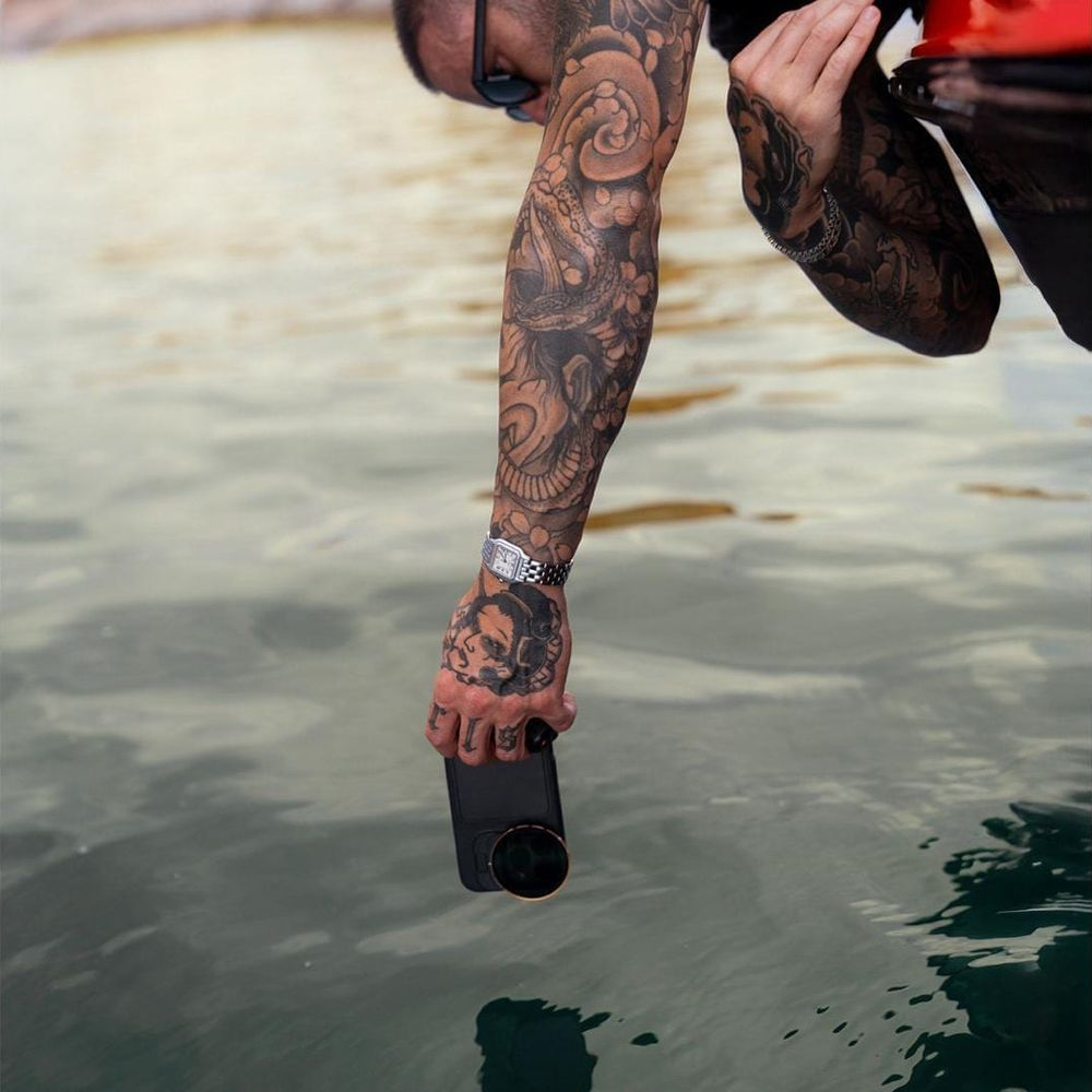 Mann mit tätowiertem Arm hält ein Handy über Wasser.