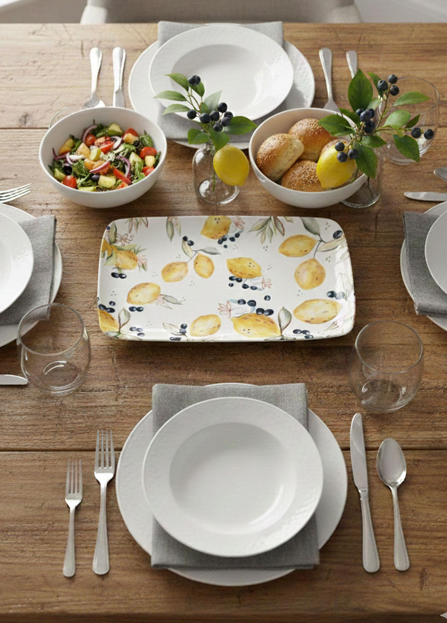 Beyaz tabaklar, çatal bıçak takımı, bardaklar, salata ve ekmek rulolarıyla donatılmış bir masa.