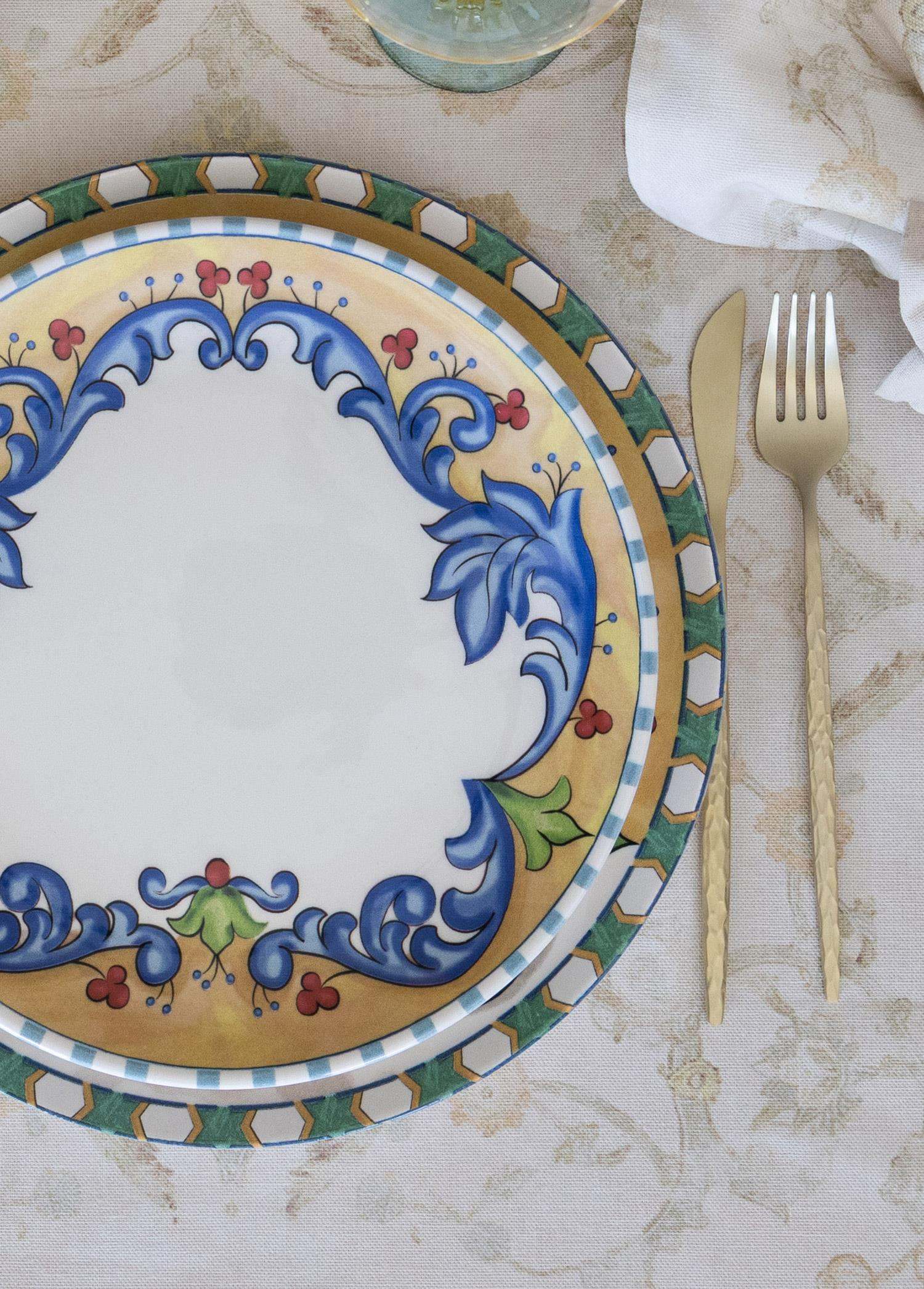 Mavi ve kırmızı çiçek desenli dekoratif bir tabak desenli masa örtüsü üzerinde duruyor. Sağda altın rengi çatal bıçak.