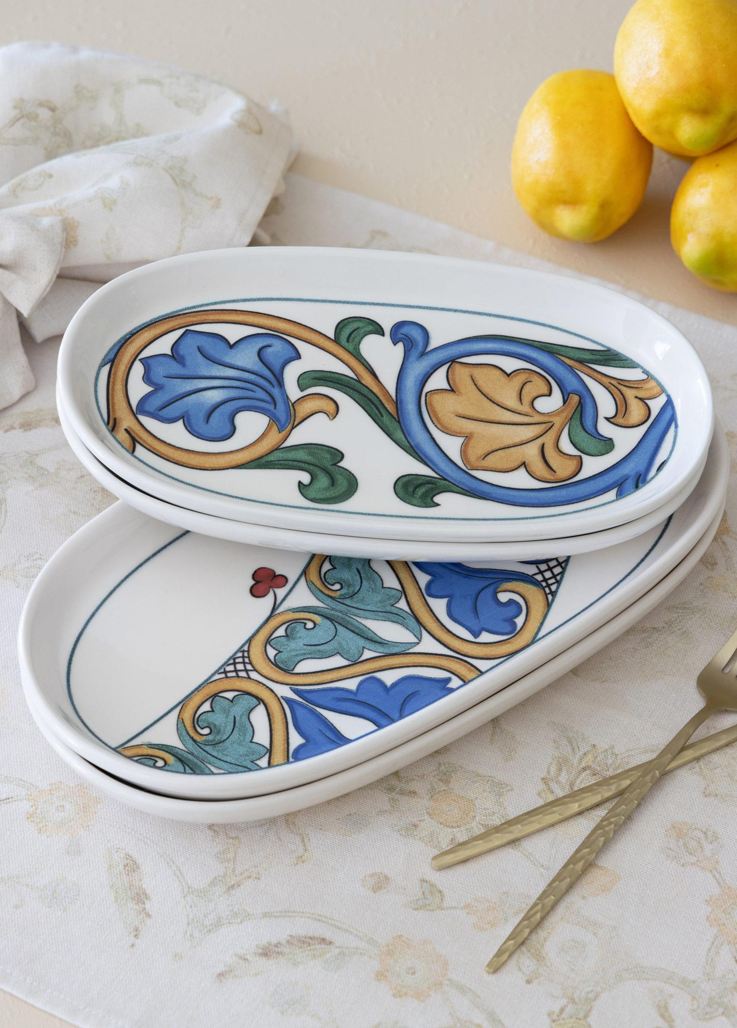 Üst üste dizilmiş, mavi, altın ve yeşil çiçek desenli iki oval tabak. Yakınlarda üç limon ve altın rengi çatallar.
