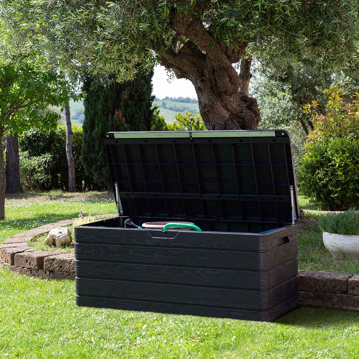 An open black storage box in a garden, containing garden tools and a hose.