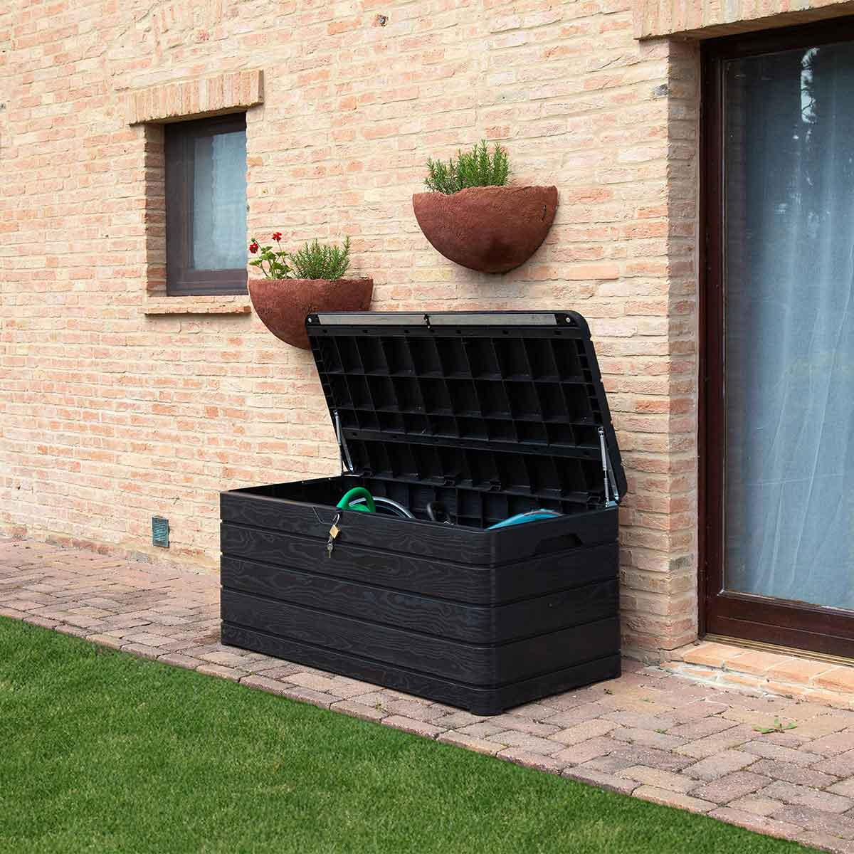 Black storage box with lid open, next to a brick wall with plants.