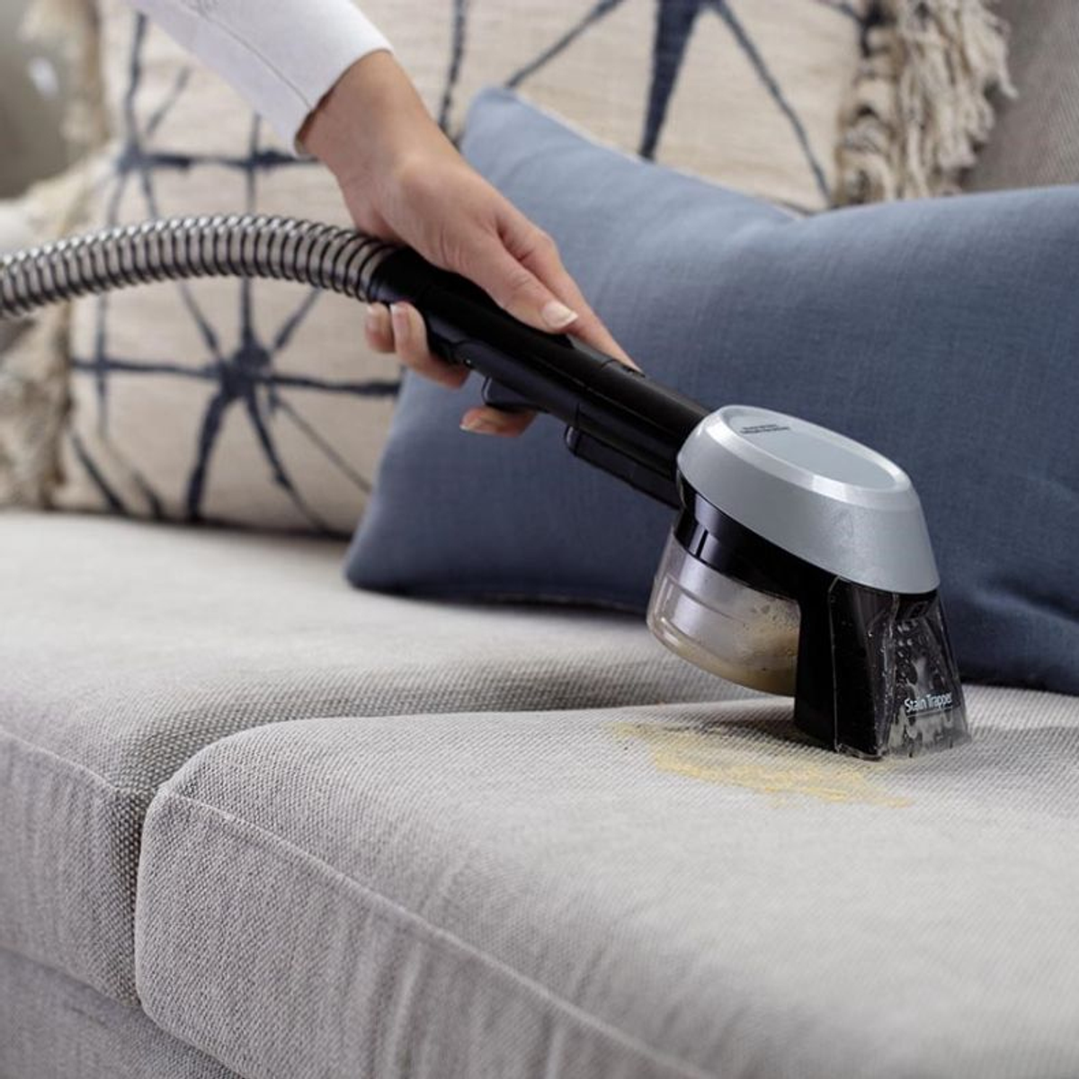 Eine Hand benutzt einen Fleckenentferner auf einem hellgrauen Sofa. Gelber Fleck sichtbar.