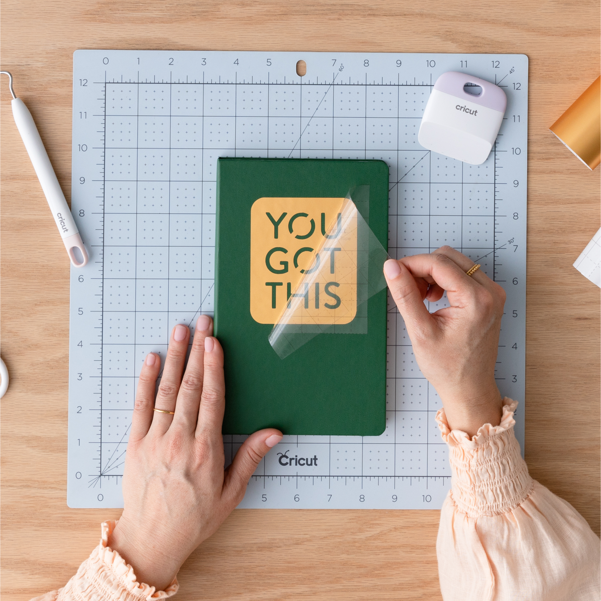 Persona aplica pegatina 'YOU GOT THIS' en cuaderno verde sobre tapete de corte.