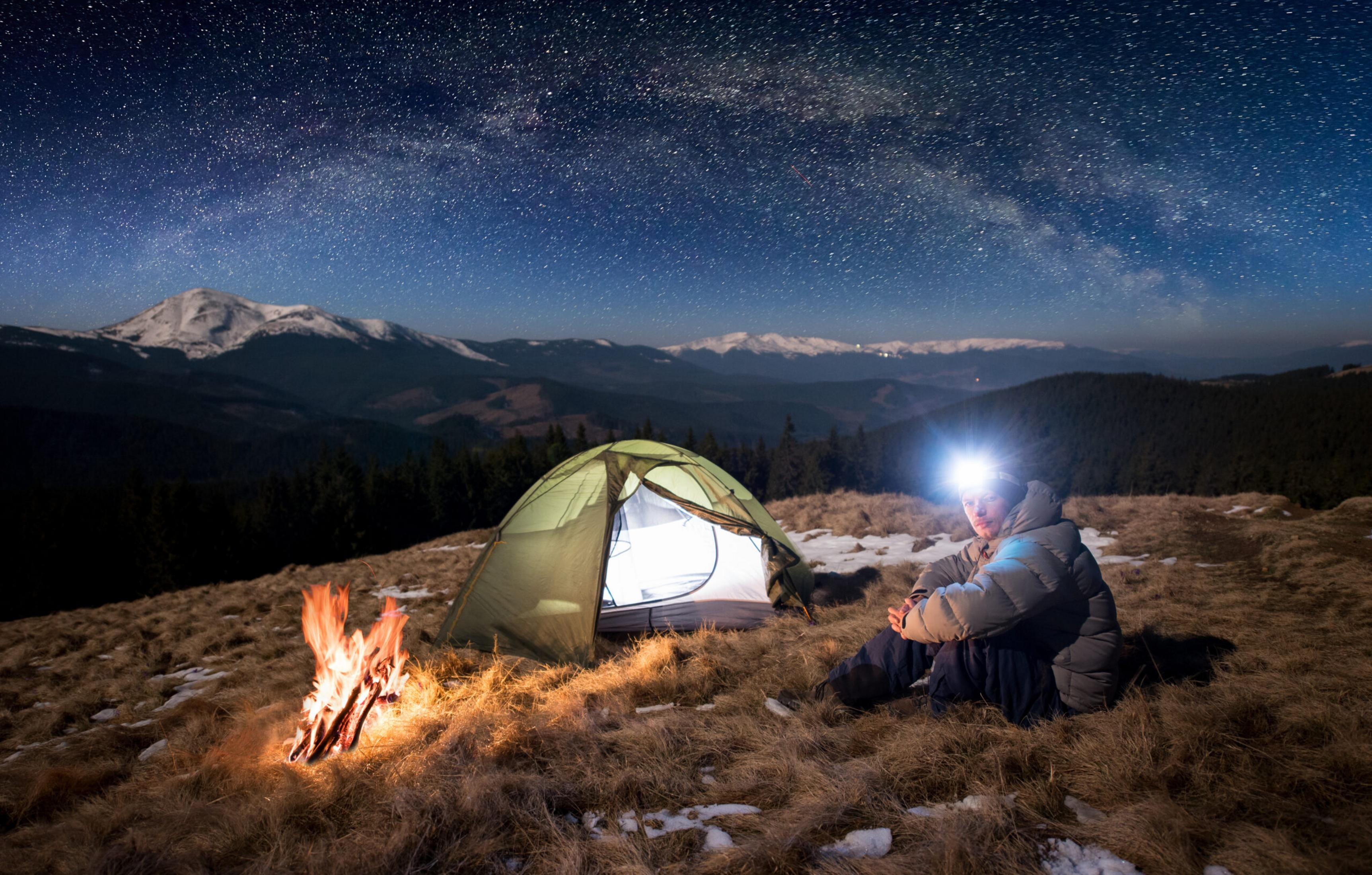 Ein Camper sitzt neben einem leuchtenden Lagerfeuer und Zelt unter einem Sternenhimmel.
