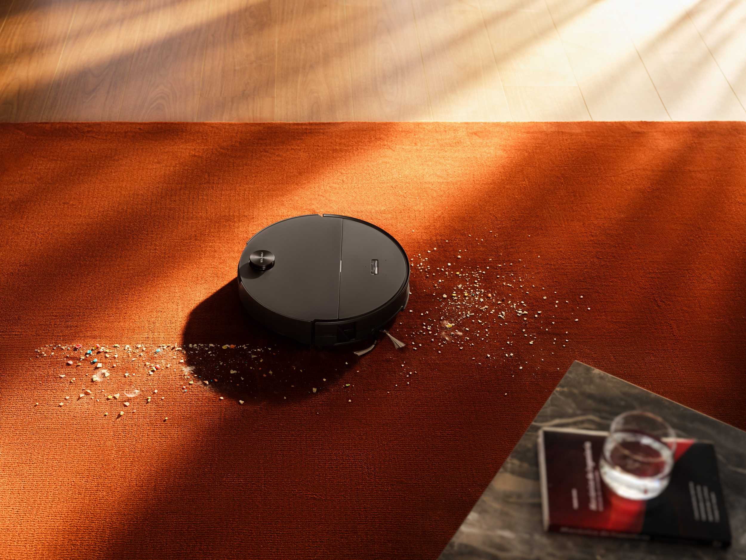 Un robot aspirateur noir nettoie des miettes d'un tapis orange.