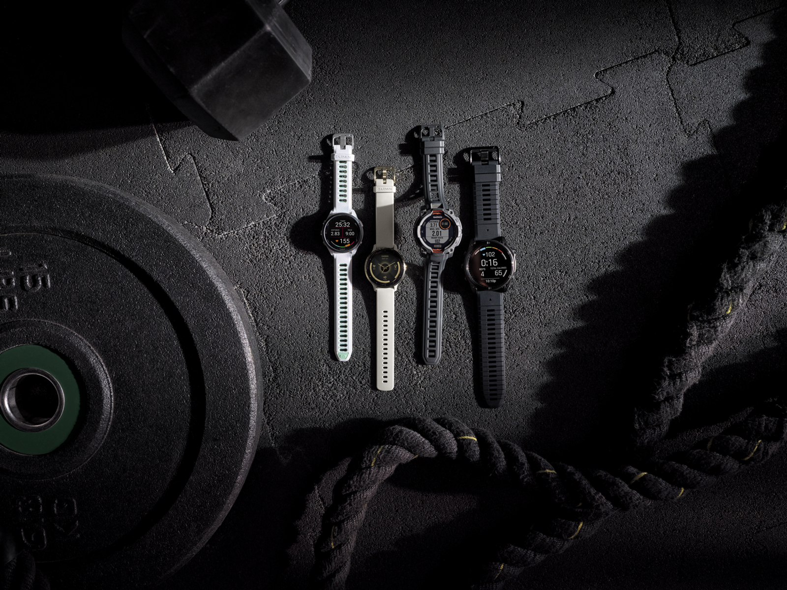 Vier Garmin horloges liggen op een donkere gymvloer naast gewichten en touwen.