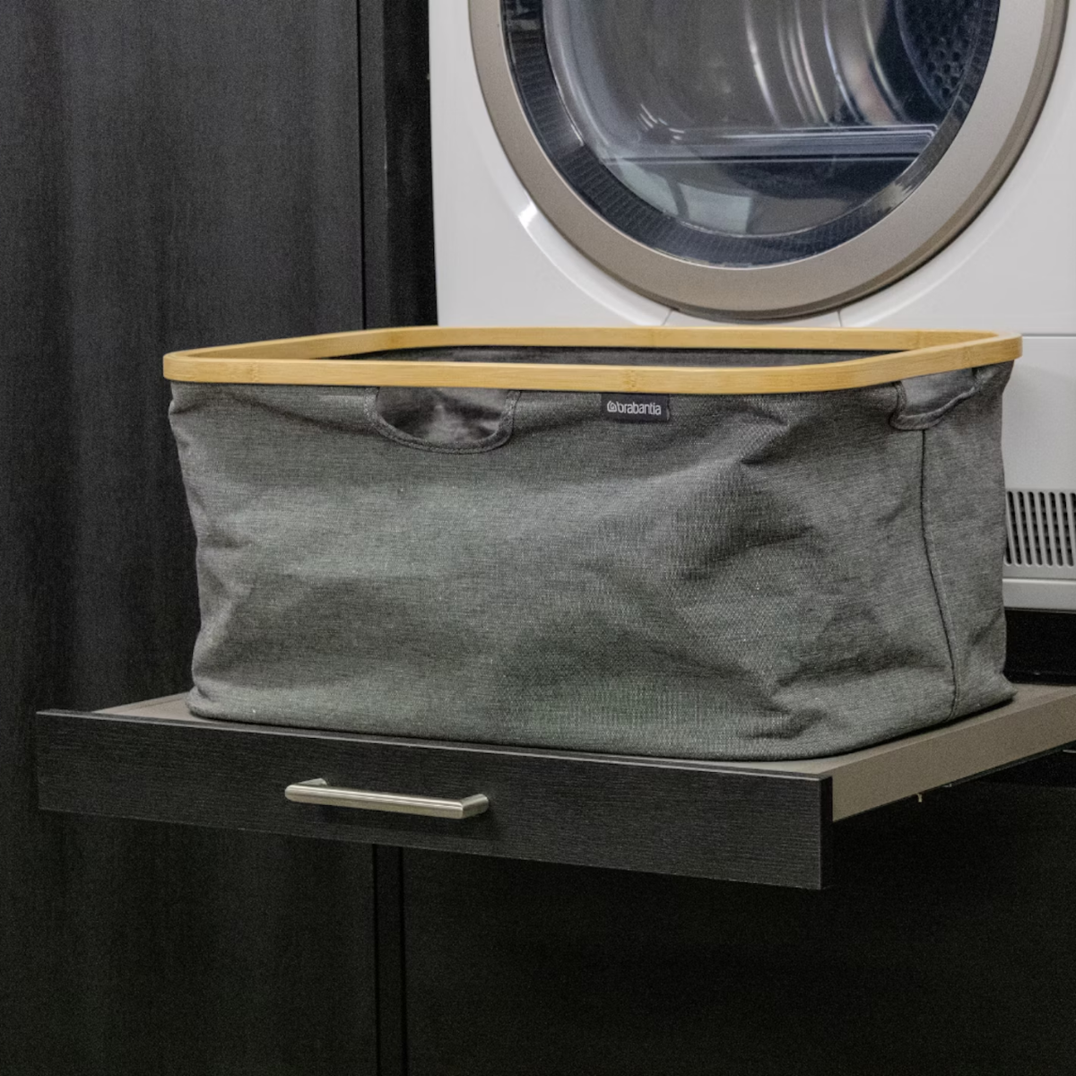 Grauer Wäschekorb mit Holzrand auf einem dunklen Regal vor einer Waschmaschine.